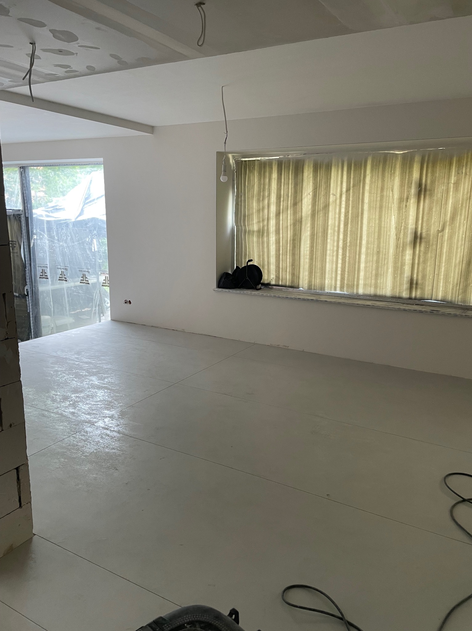 Wnętrze w trakcie remontu: gładkie, jasne ściany, wylewka na podłodze, sufit z płyt karton-gips, okno z zasłoną. Widoczne narzędzia i materiały budowlane.