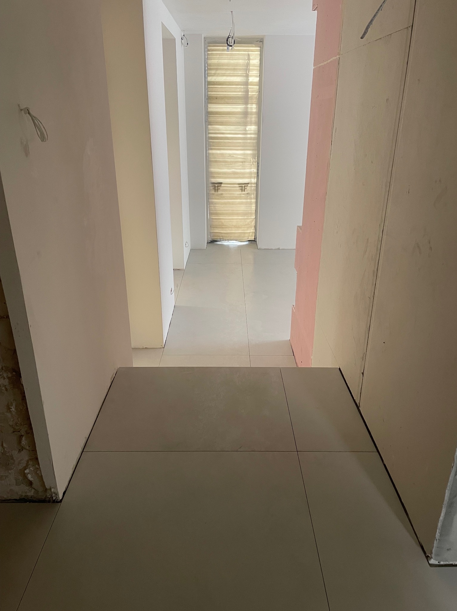 Wnętrze w trakcie remontu: świeżo położona, duża, szara płytka podłogowa, surowe ściany w odcieniach beżu i różu, widoczne kable i elementy instalacji.