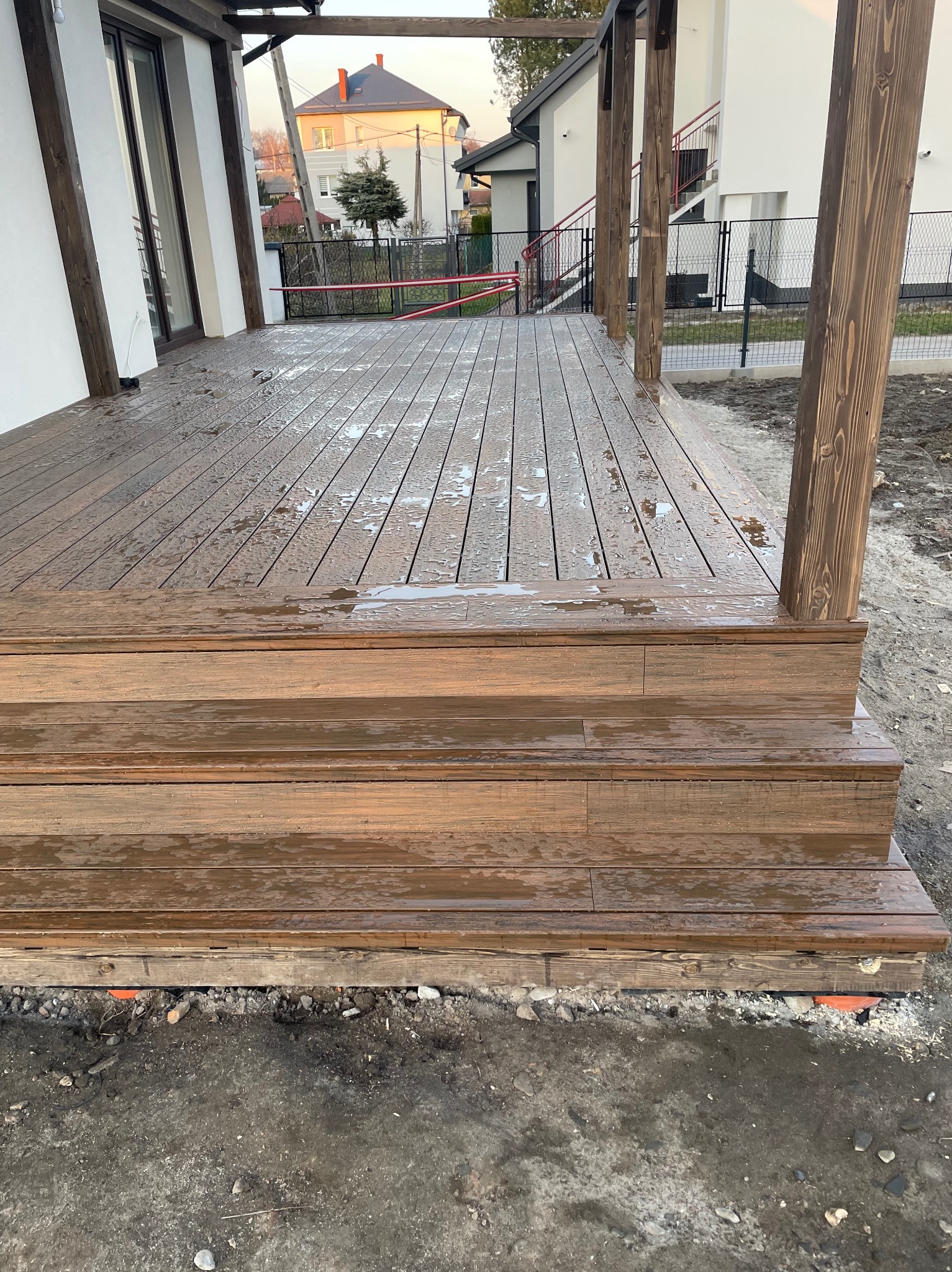 Drewniany taras z trzema stopniami, po deszczu. Widoczne krople wody na deskach i elementy konstrukcyjne z ciemnego drewna. W tle budynki mieszkalne.