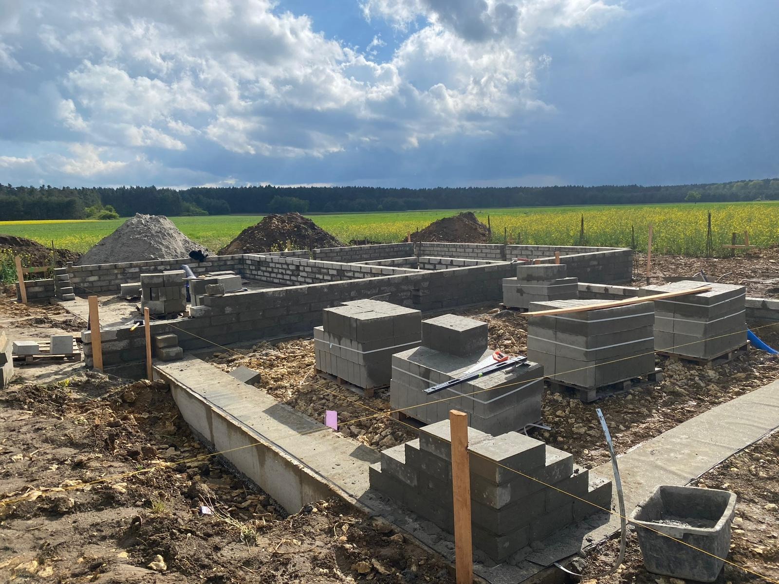 Budowa domu: fundamenty i pierwsze warstwy ścian z bloczków betonowych na tle zielonego pola i pochmurnego nieba. Widoczne narzędzia i materiały budowlane.