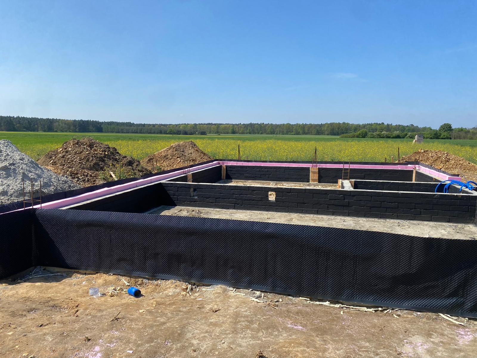 Fundamenty domu z czarną izolacją i różowym ociepleniem na tle zielonego pola i błękitnego nieba. Widoczne hałdy ziemi i piasku.