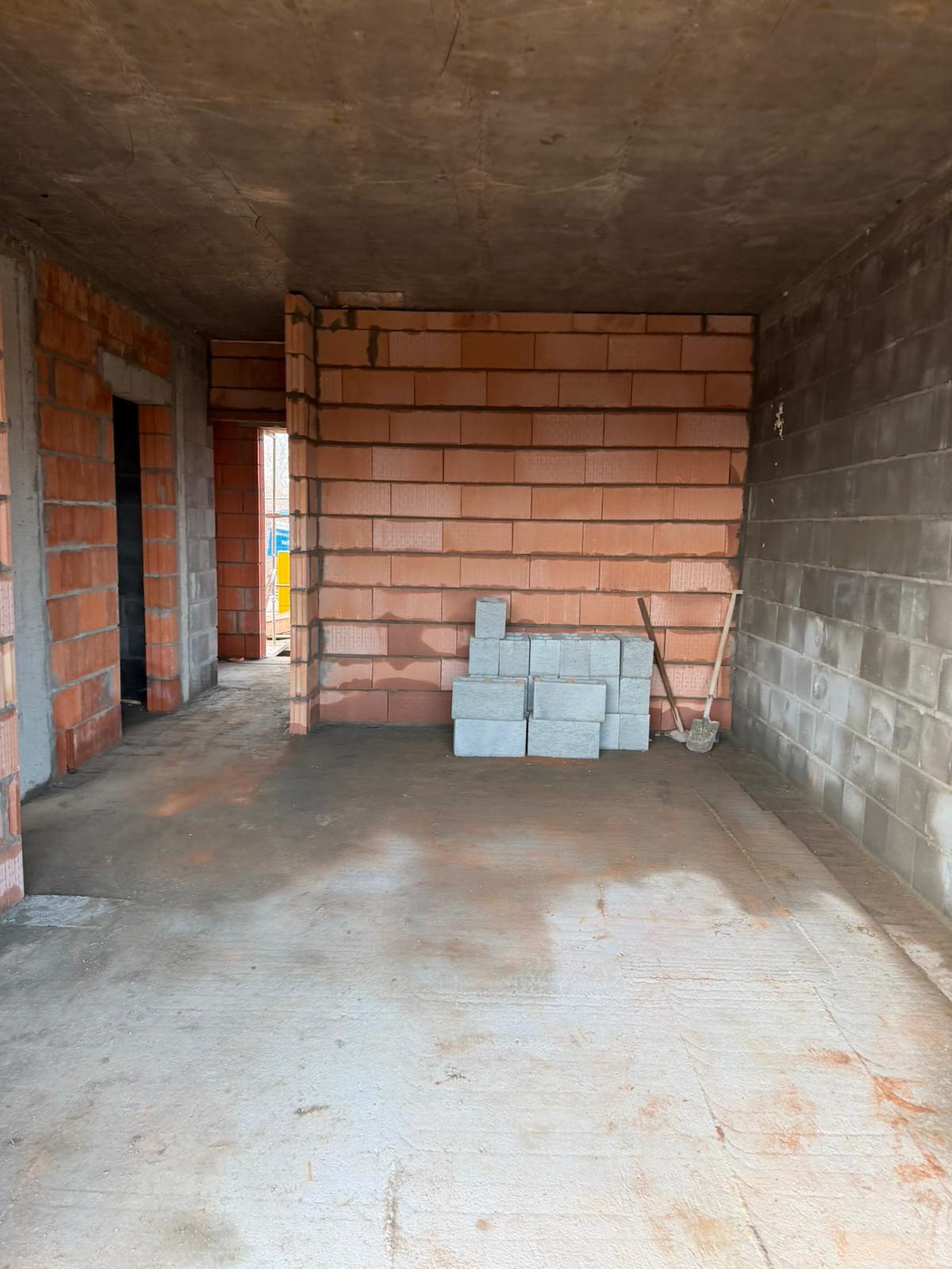 Wnętrze w trakcie budowy: ściany z czerwonej cegły, szare bloczki, betonowa podłoga i sufit. Widoczna łopata. Surowy stan, brak wykończenia.