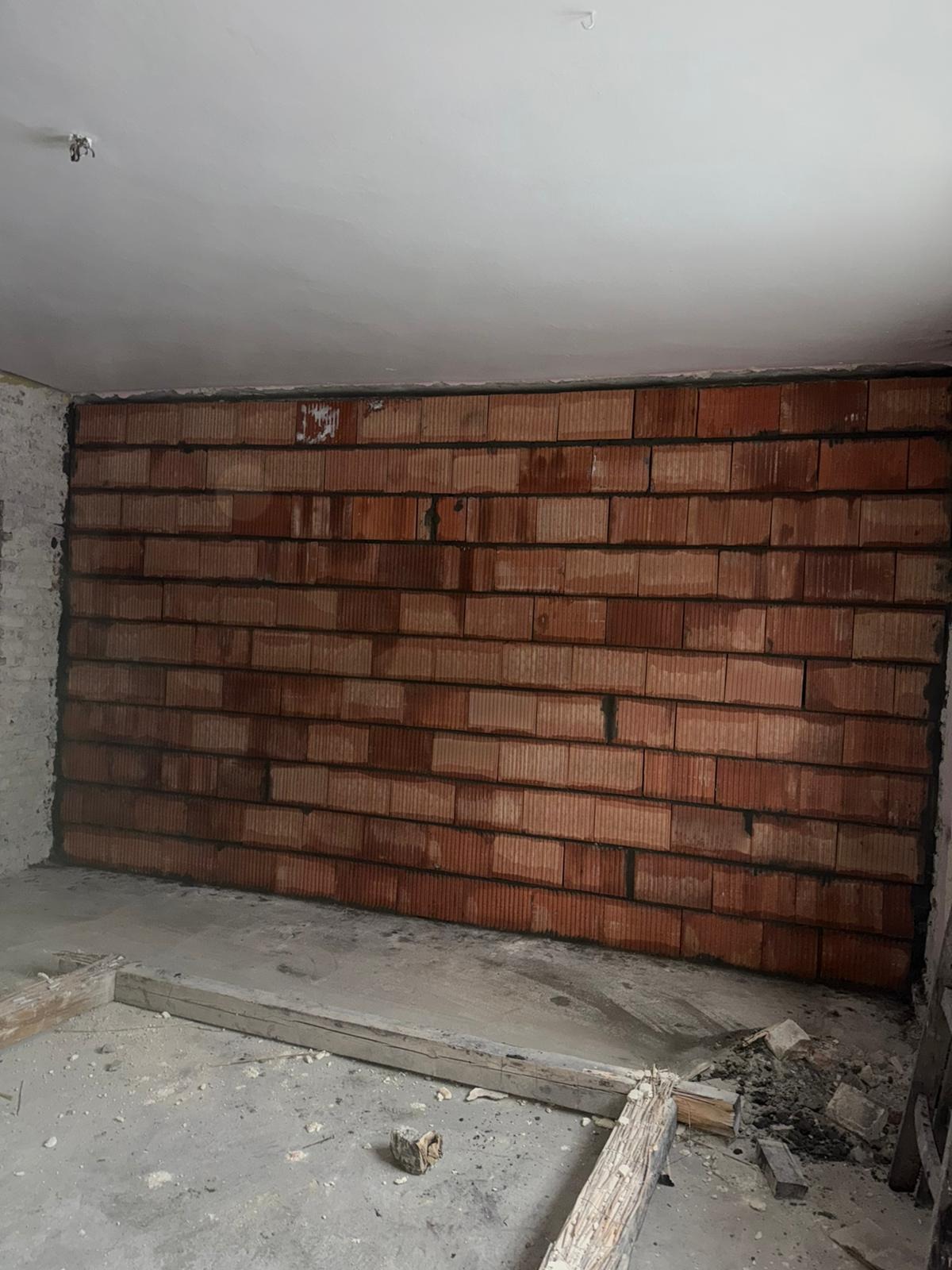 Ściana z czerwonej cegły dziurawki w surowym wnętrzu, widoczne ślady zaprawy, fragment podłogi z resztkami gruzu i drewnianych elementów szalunku.