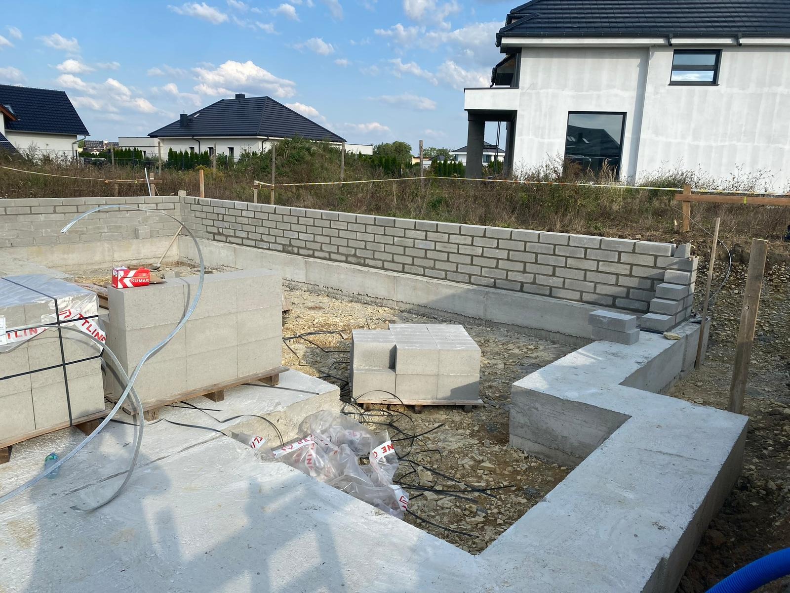 Wznoszenie muru z bloczków betonowych na fundamencie domu jednorodzinnego. Widoczne palety z bloczkami i częściowo wymurowana ściana. W tle dom w stanie surowym otwartym.