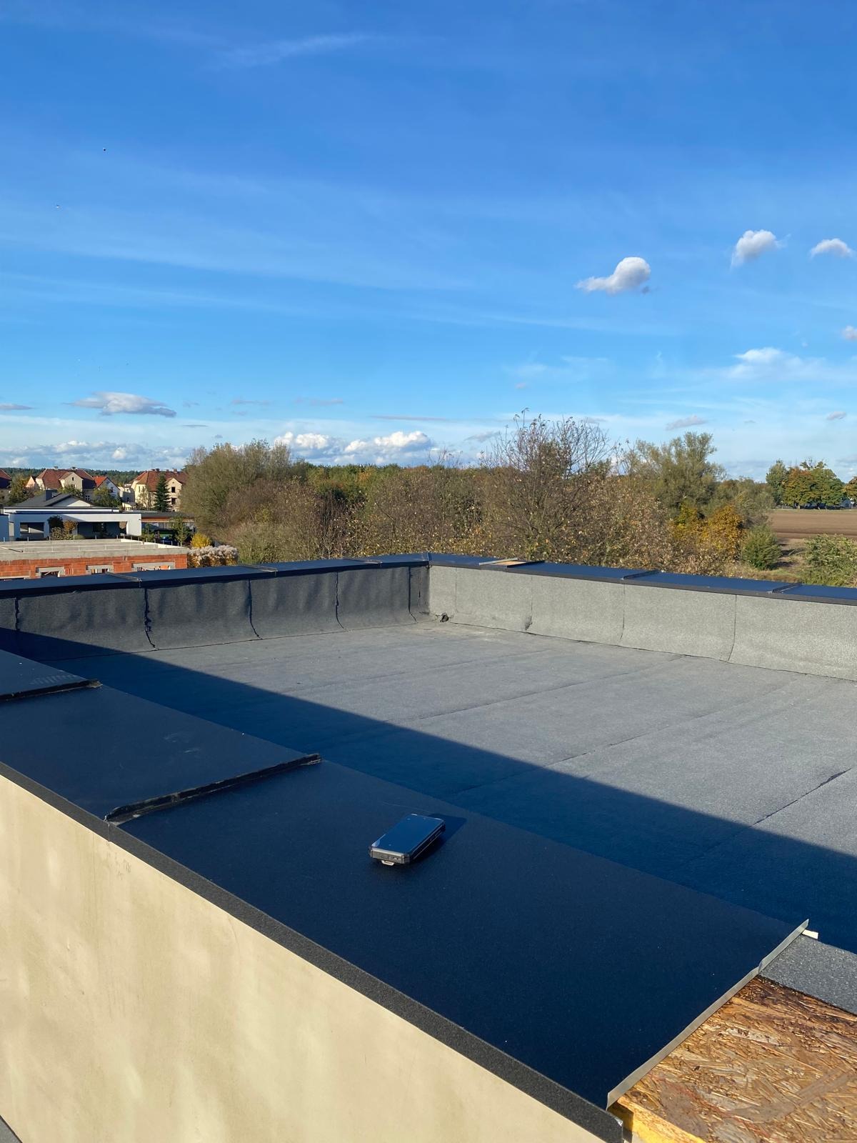 Wykończony płaski dach pokryty papą termozgrzewalną, z widocznym smartfonem na krawędzi. W tle błękitne niebo i zabudowa jednorodzinna.