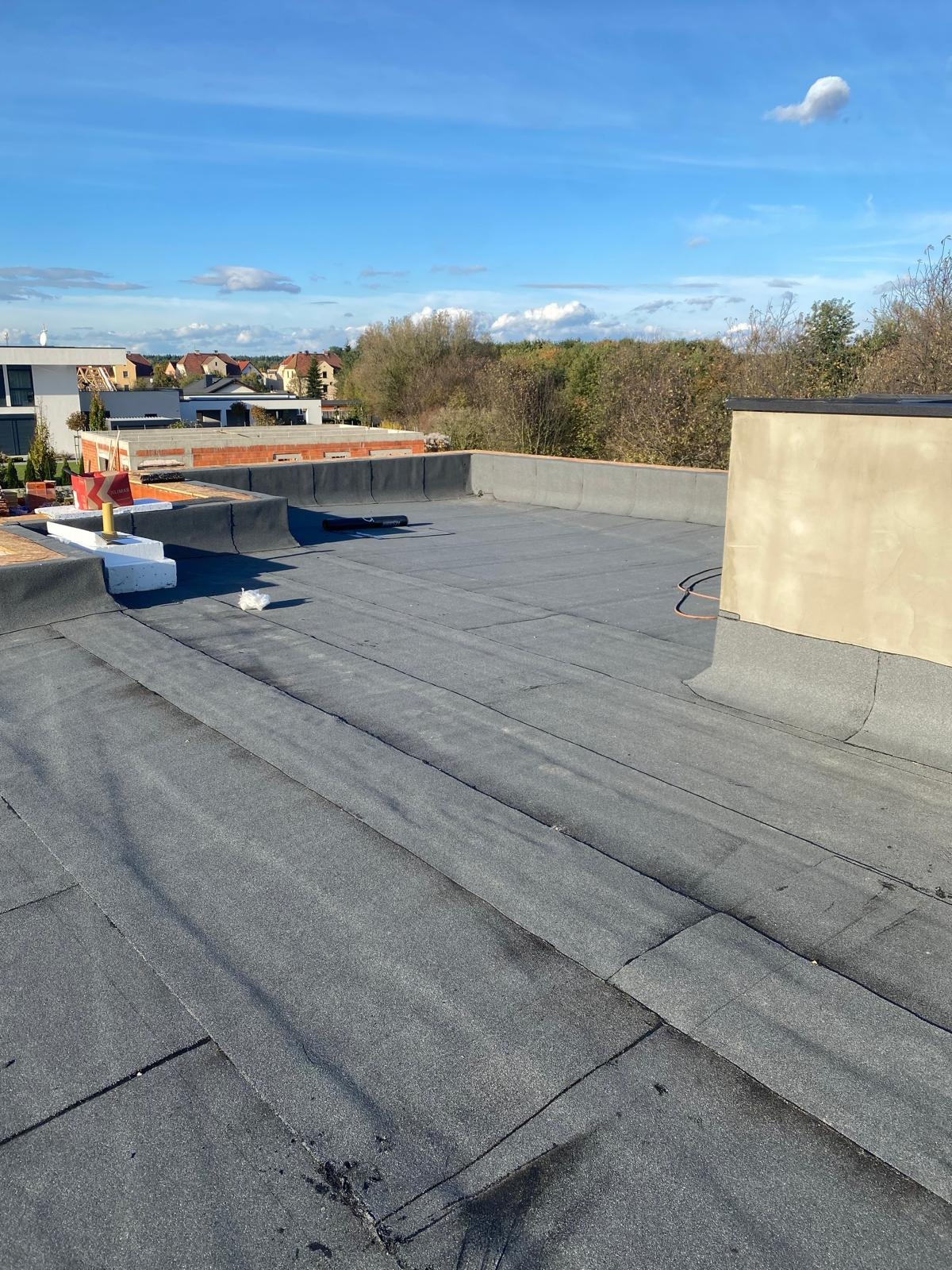 Wykończony płaski dach pokryty papą termozgrzewalną, widok z góry na tle zabudowy jednorodzinnej i błękitnego nieba z chmurami. Detal łączeń papy.