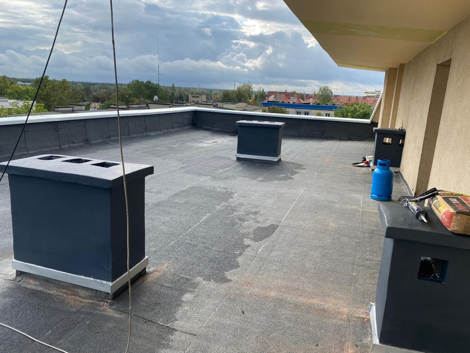 Wykończony dach płaski z kominami wentylacyjnymi, widoczne narzędzia i materiały budowlane. W tle zabudowa miejska i pochmurne niebo.