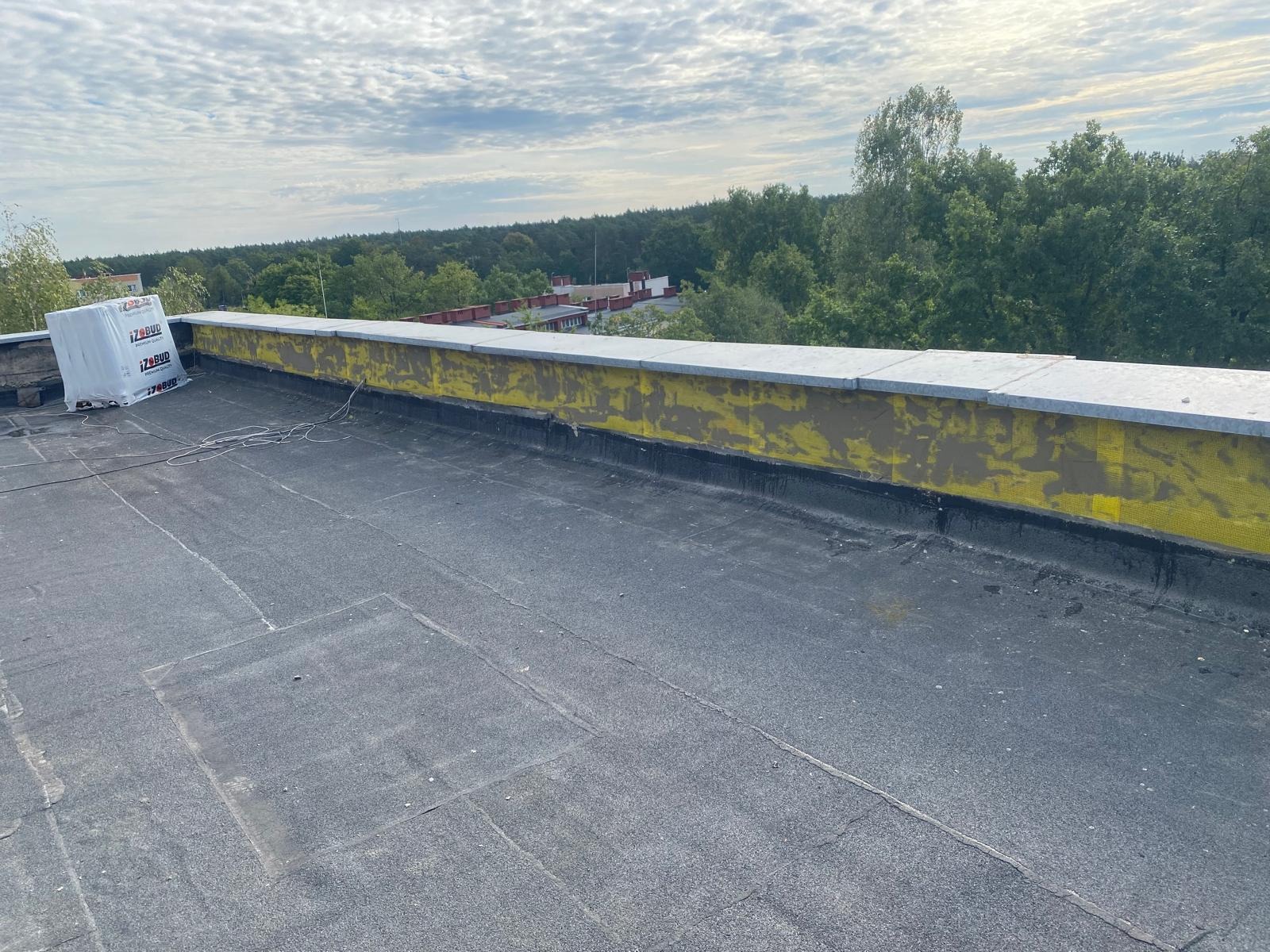 Płaski dach pokryty papą, z widoczną izolacją termiczną ściany attykowej i częściowo zabezpieczonym materiałem budowlanym. W tle zieleń drzew i budynki.