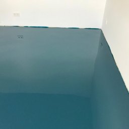 BLAN-BUD Marek Gutowski - Błękitna, błyszcząca posadzka żywiczna w rogu pomieszczenia, zabezpieczona taśmą malarską przy ścianach. Widoczne gniazdka elektryczne na ścianie.