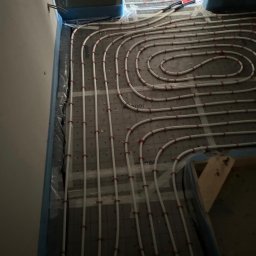 BLAN-BUD - Białe rury ogrzewania podłogowego, przymocowane czerwonymi uchwytami do szarej maty izolacyjnej, ułożone spiralnie w pomieszczeniu przed wylaniem posadzki.