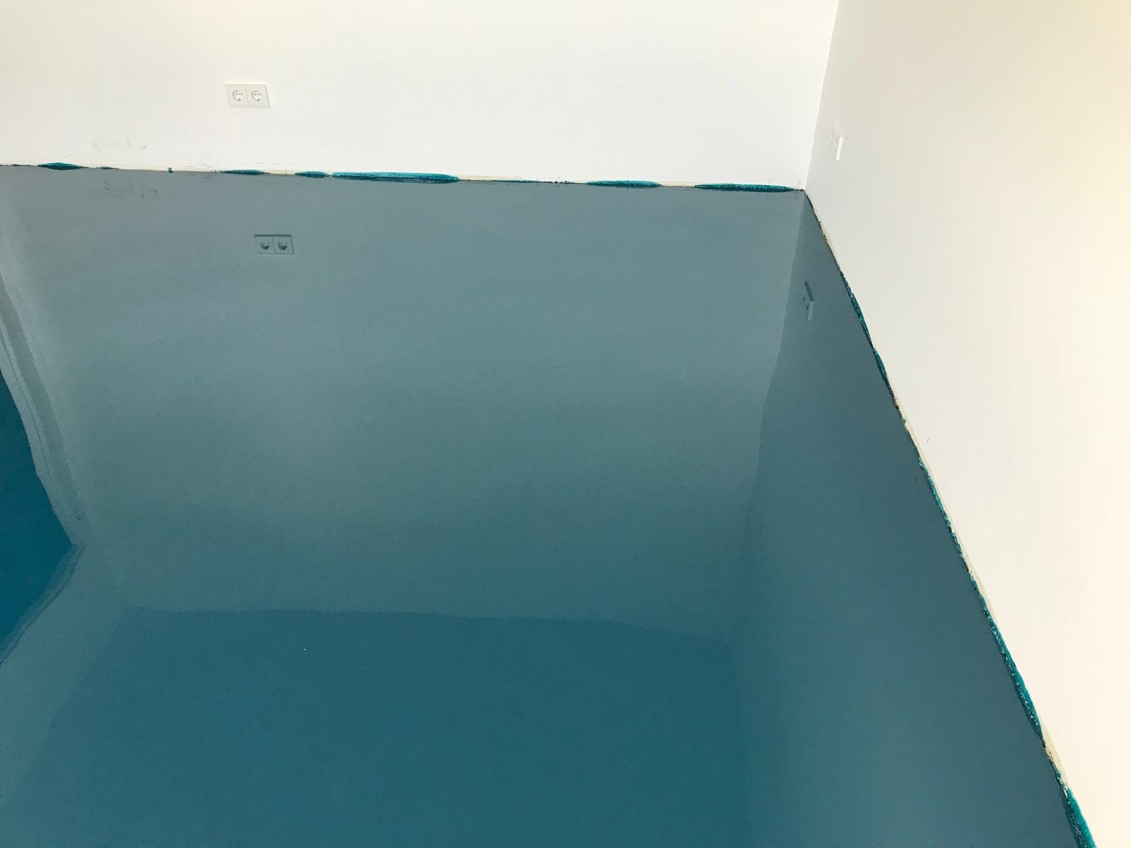 Błękitna, błyszcząca posadzka żywiczna w rogu pomieszczenia, zabezpieczona taśmą malarską przy ścianach. Widoczne gniazdka elektryczne na ścianie.