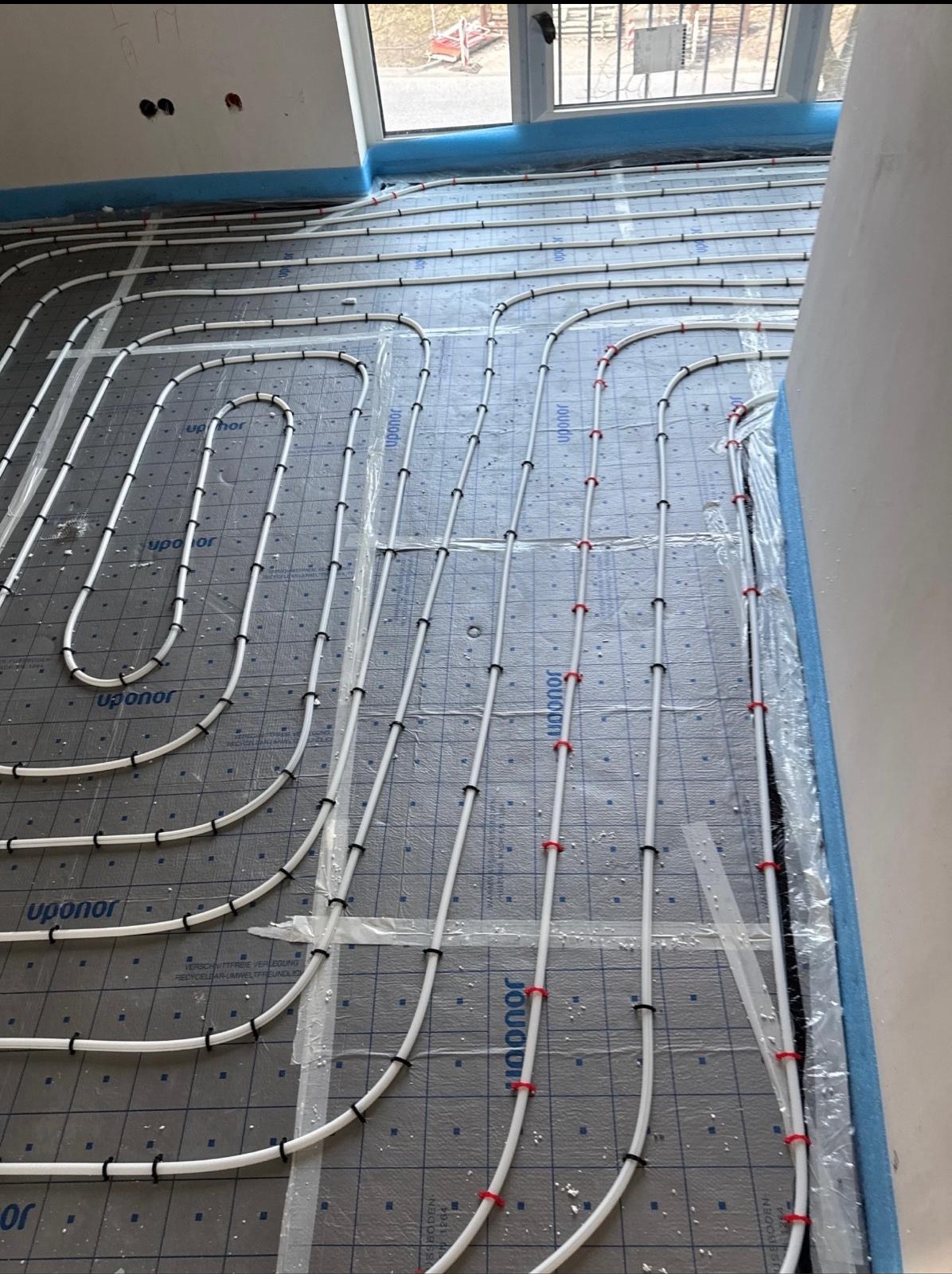 Białe rury ogrzewania podłogowego marki Uponor, ułożone na szarej macie izolacyjnej z niebieską taśmą brzegową, widoczne przez okno. Instalacja w trakcie montażu.