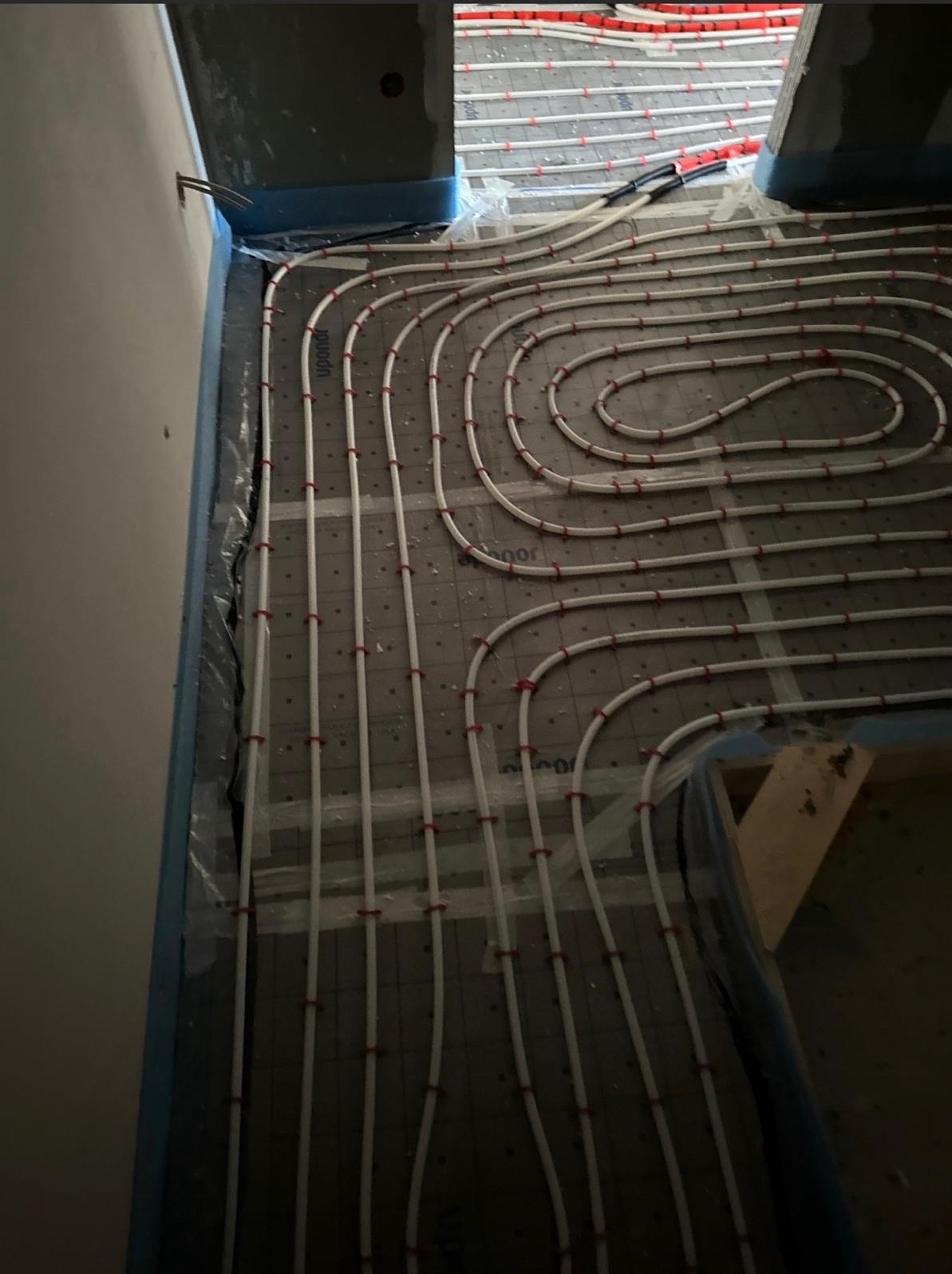 Białe rury ogrzewania podłogowego, przymocowane czerwonymi uchwytami do szarej maty izolacyjnej, ułożone spiralnie w pomieszczeniu przed wylaniem posadzki.