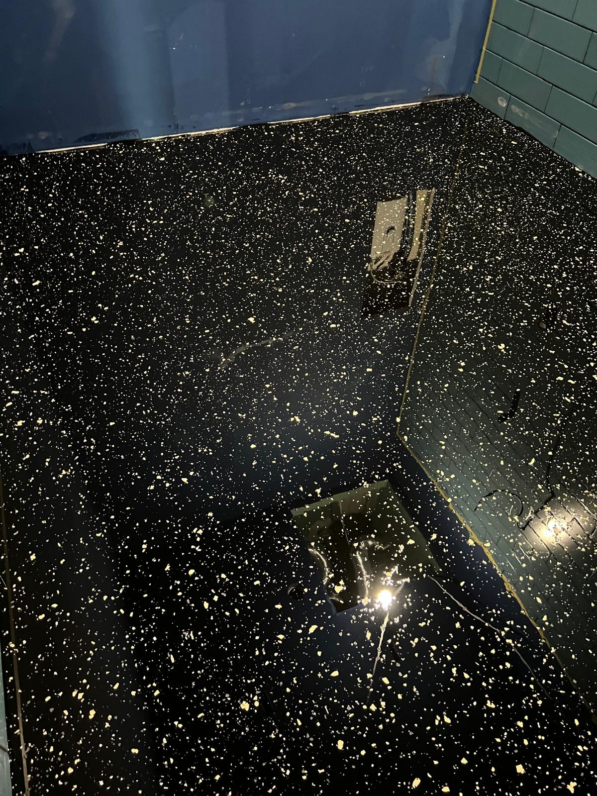 Błyszcząca, czarna posadzka żywiczna z drobnymi, złotymi plamkami, odbijająca światło. Widoczny fragment ściany z niebieskimi płytkami.