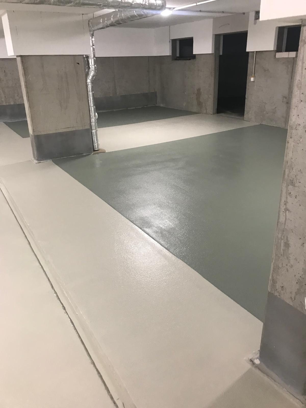 Gładka, dwukolorowa posadzka w odcieniach szarości i beżu w surowym wnętrzu. Widoczne betonowe filary i fragment instalacji wentylacyjnej. Minimalistyczny design.