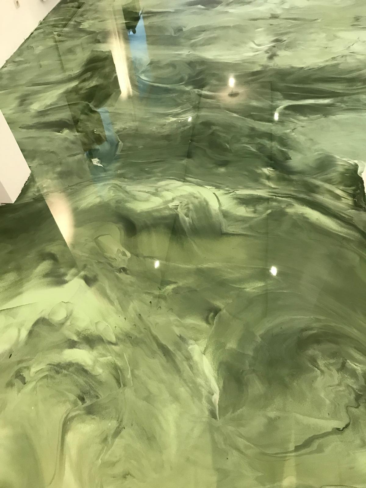 Błyszcząca, zielona posadzka z efektem marmuru, wykonana z żywicy. Widoczne refleksy światła na gładkiej powierzchni. Fragment ściany w lewym górnym rogu.