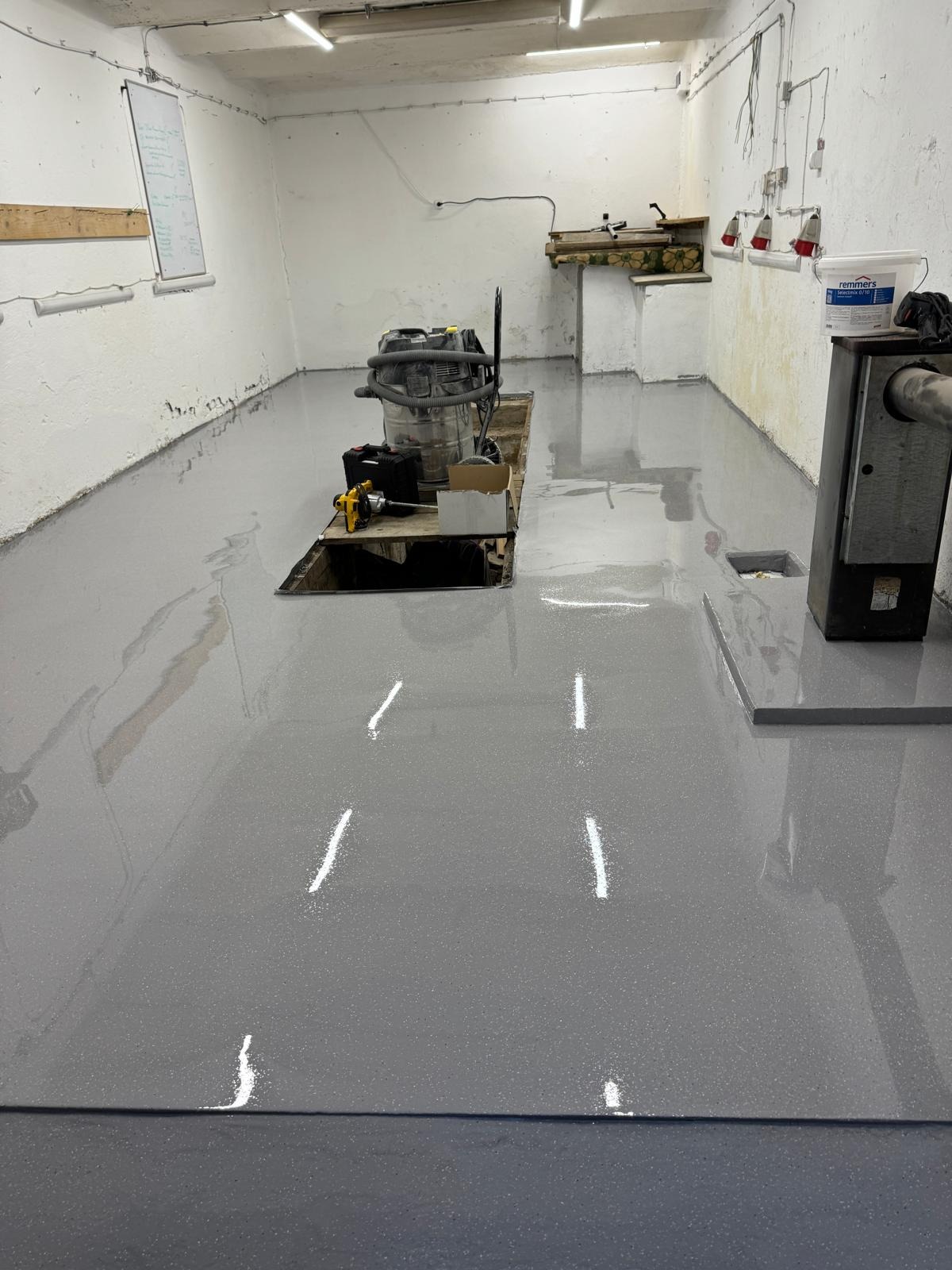 Szara, błyszcząca posadzka w pomieszczeniu gospodarczym z narzędziami i sprzętem budowlanym. Widoczne ślady po białym proszku na powierzchni.