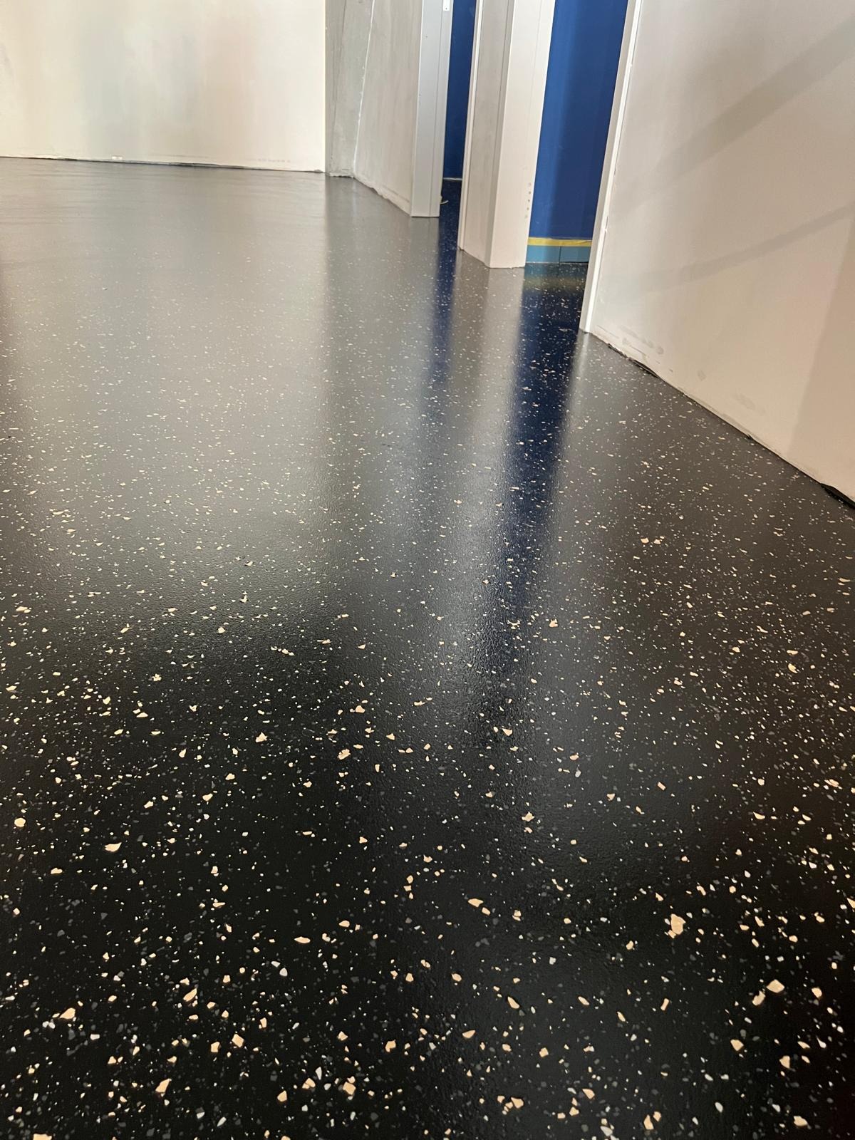 Błyszcząca, czarna posadzka z beżowymi drobinkami, odbijająca światło. Widoczne białe ściany i fragment niebieskiej kolumny w tle. Gładka powierzchnia, perspektywa z dołu.