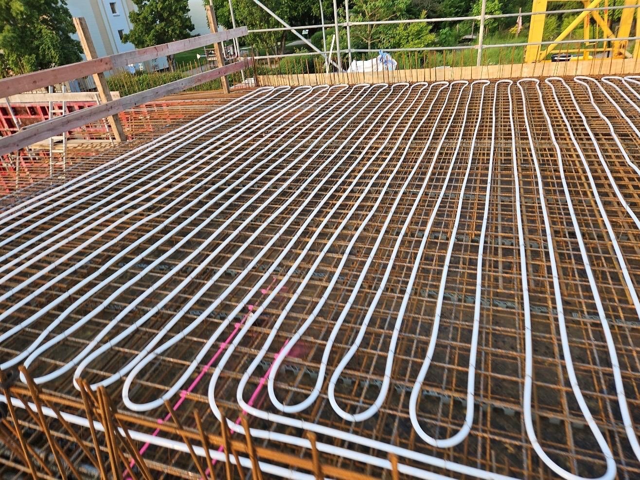 Białe rury ogrzewania podłogowego ułożone równolegle na siatce zbrojeniowej, przygotowane do zalania posadzką. Widok z góry, częściowo w cieniu.