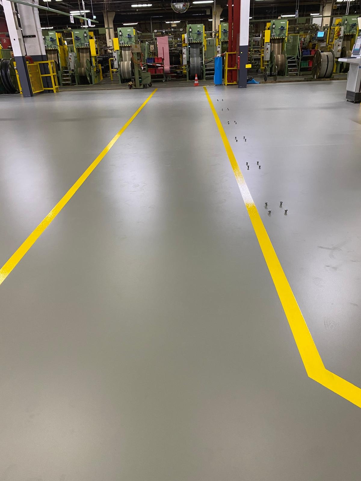 Szara posadzka przemysłowa z żółtymi liniami wyznaczającymi ciągi komunikacyjne w hali produkcyjnej. Widoczne śruby na podłodze.