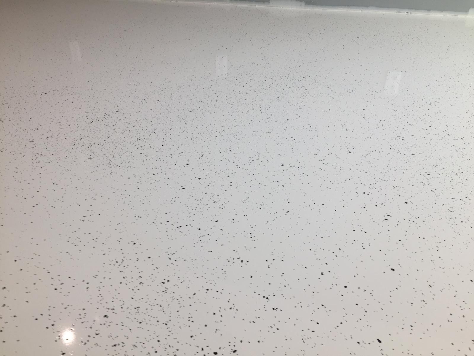 Jasna, gładka posadzka żywiczna z drobnymi, czarnymi inkluzjami, tworzącymi subtelny wzór. Widoczne delikatne refleksy światła na powierzchni.