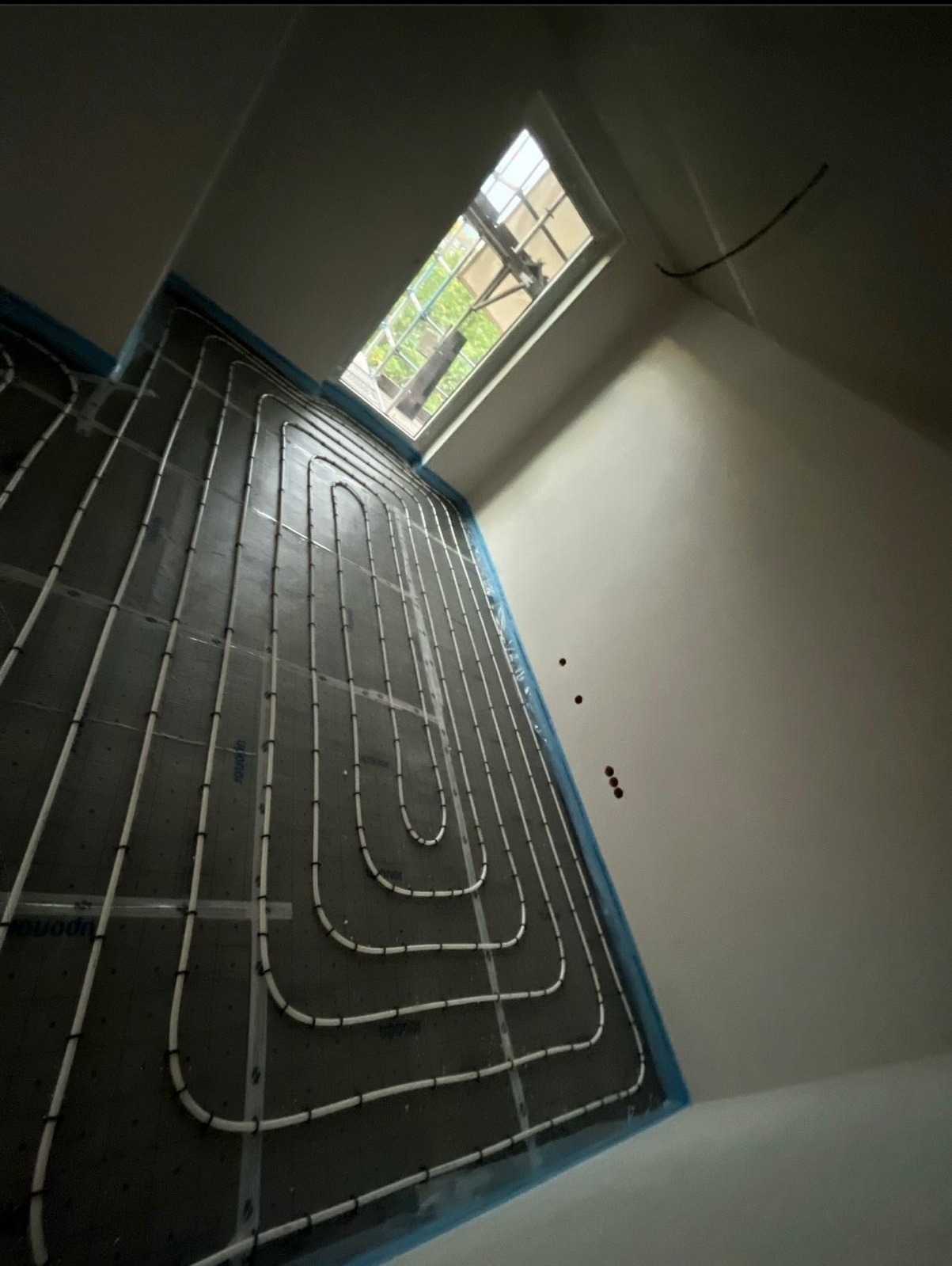 Białe rury ogrzewania podłogowego ułożone na czarnej macie izolacyjnej, widok z góry, okno dachowe w tle, niebieska folia zabezpieczająca przy ścianach.