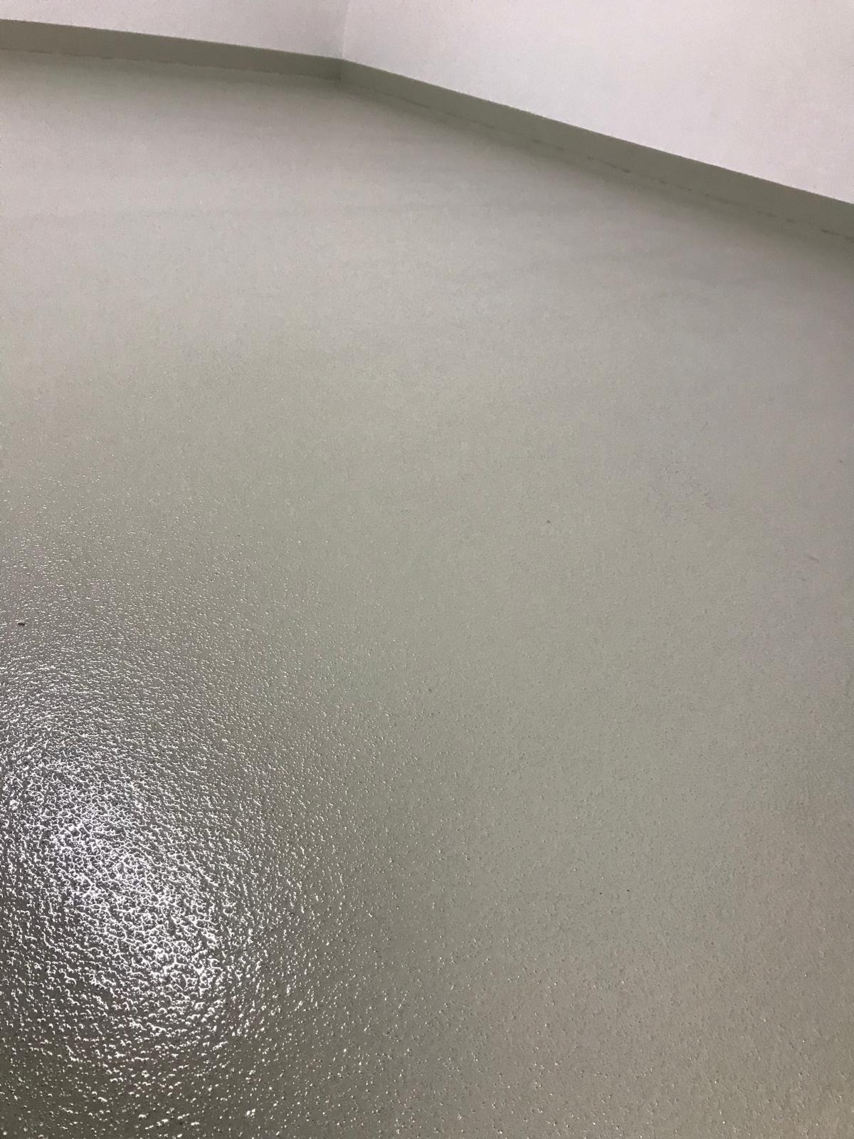 Jasnoszara, gładka posadzka z delikatnym połyskiem, widoczna przy ścianie. Minimalistyczne wykończenie, jednolite tło, brak elementów dekoracyjnych.