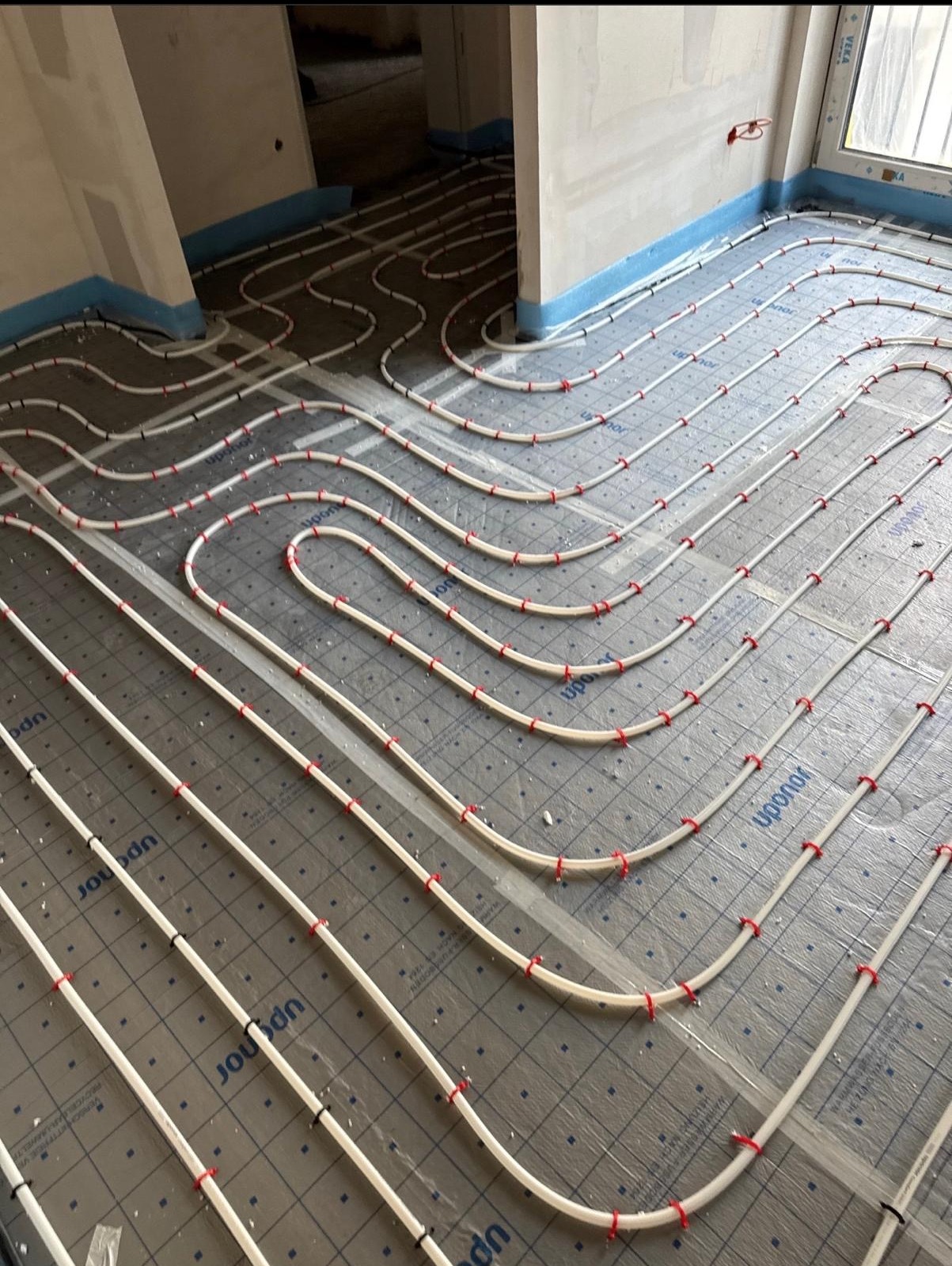 Białe rury ogrzewania podłogowego Uponor, mocowane czerwonymi klipsami na szarej folii z nadrukiem, ułożone w pętle na przygotowanym podłożu w nowym budynku.