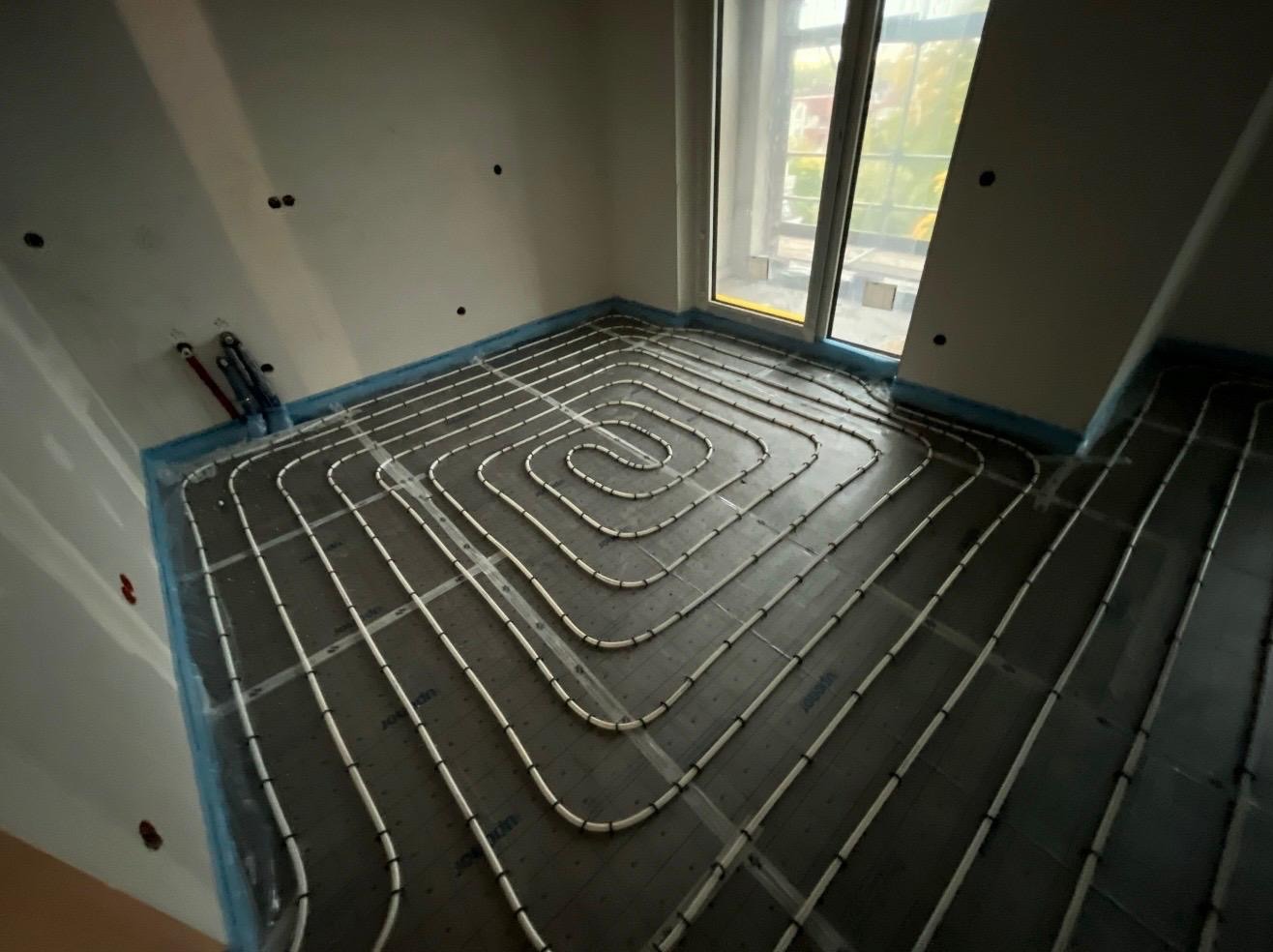 Białe rury ogrzewania podłogowego ułożone spiralnie na czarnej macie w jasnym pomieszczeniu z oknem. Widoczne przyłącza rur na ścianie.