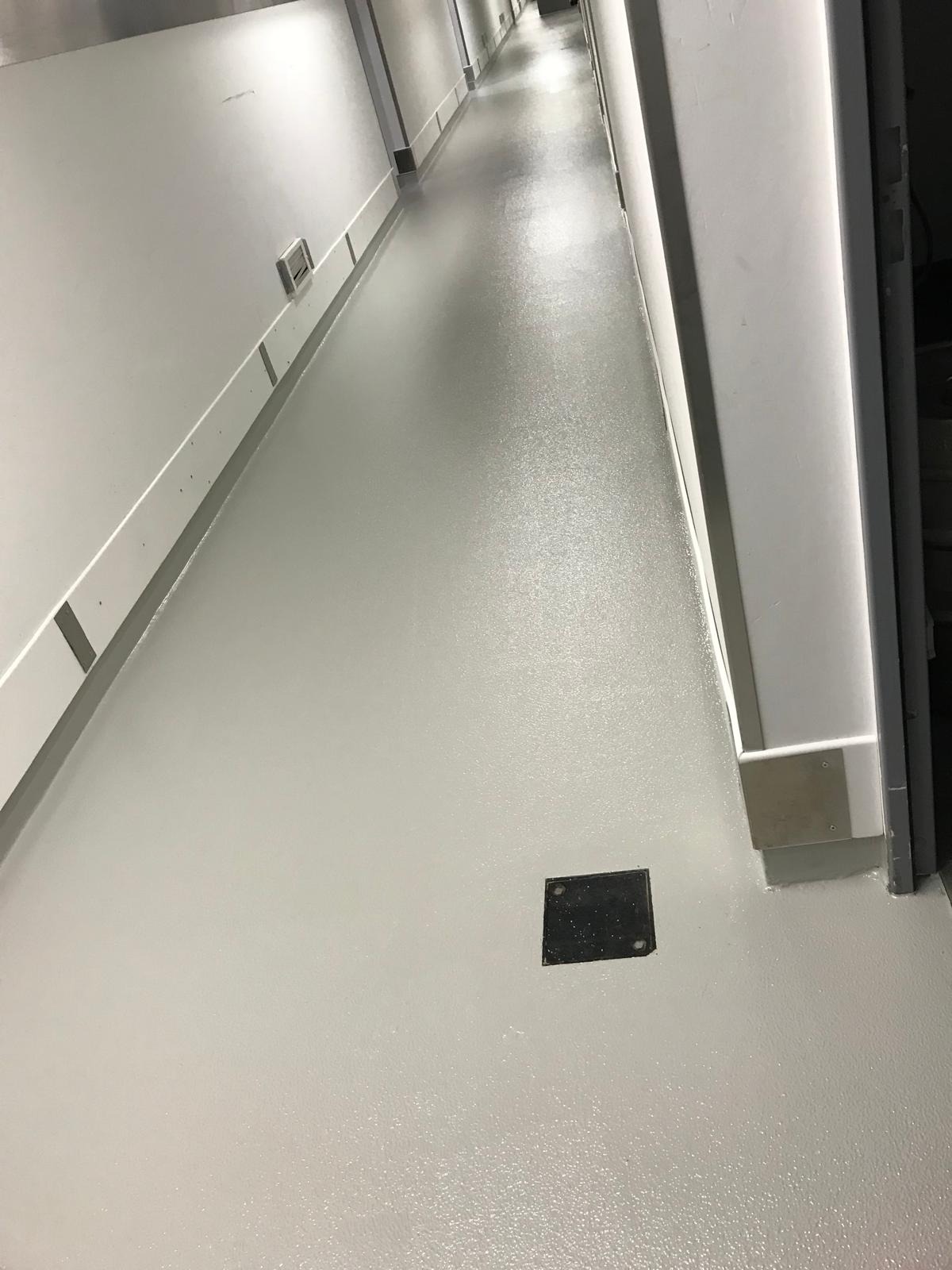 Jasnoszara, błyszcząca posadzka żywiczna w długim korytarzu z białymi ścianami i czarną kratką ściekową. Perspektywa z głębi korytarza.