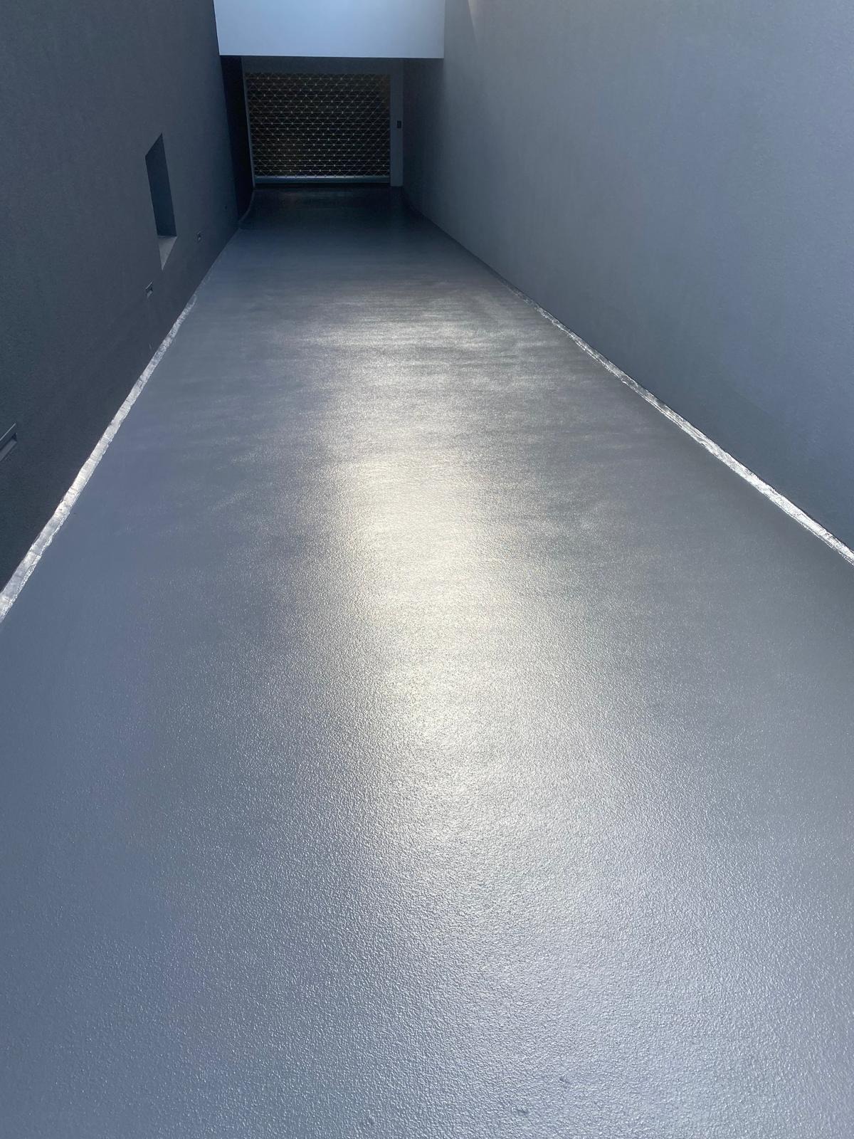 Gładka, szara posadzka w garażu podziemnym, wykonana z żywicy, z widocznym odpływem liniowym przy ścianie. Metalowa brama garażowa w tle.