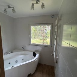 Andrei Sotnik - Biała wanna z hydromasażem w narożnej łazience z oknem na zieleń. Na ścianach jasne płytki, na podłodze panele imitujące drewno. Nowoczesne oświetlenie sufitowe.