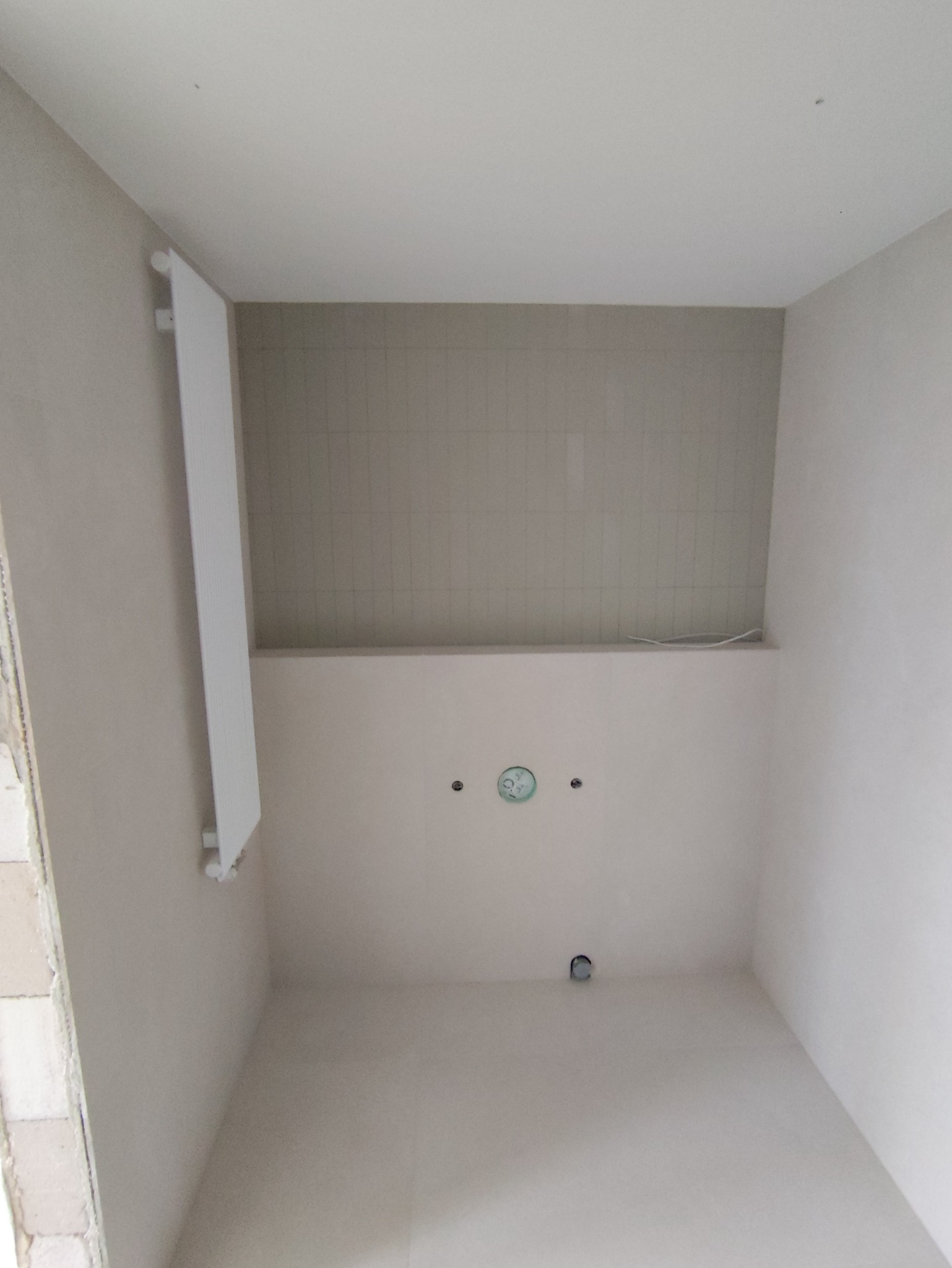 Wnętrze łazienki w trakcie remontu: widoczne płytki na ścianie, grzejnik drabinkowy, przygotowane punkty instalacyjne. Ściany w jasnych odcieniach, surowy stan wykończenia.