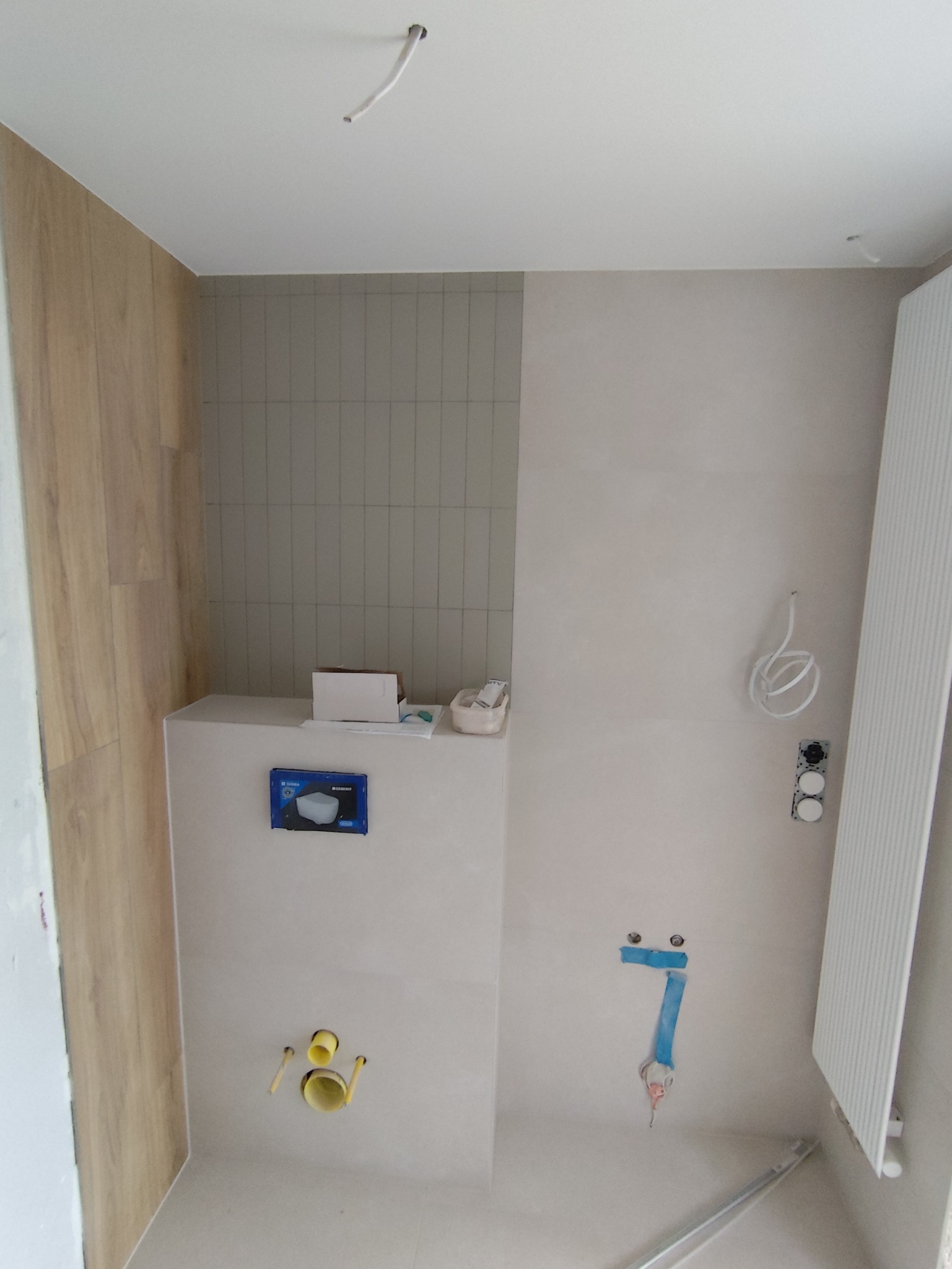 Wnętrze łazienki w trakcie wykończenia: częściowo położone płytki, zamontowany stelaż podtynkowy WC, widoczne instalacje wod-kan i elektryczne, grzejnik.