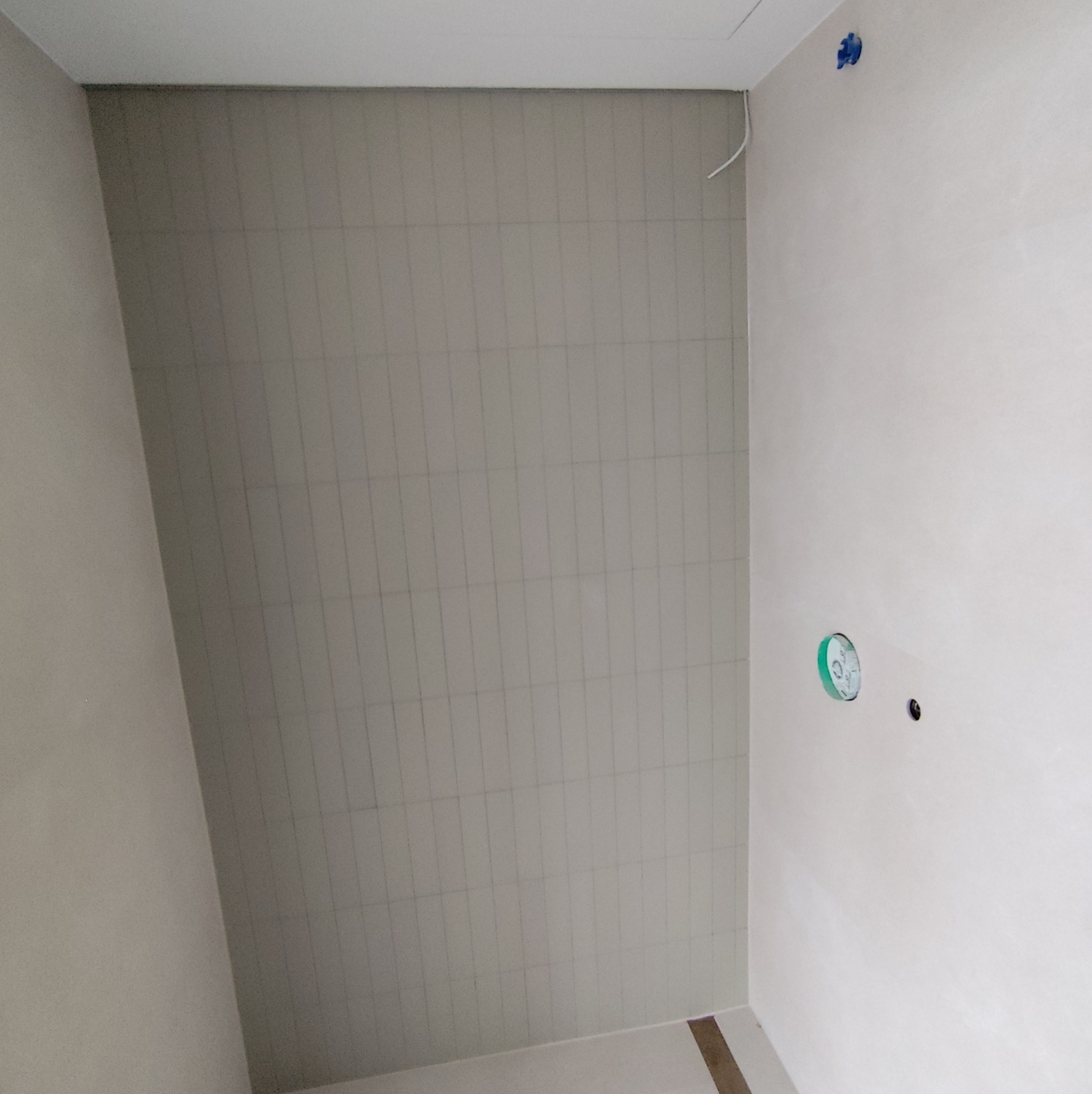 Ściana łazienki z ułożonymi jasnymi płytkami w pionowym układzie, widoczne przygotowane punkty instalacji elektrycznej i wodnej, zabezpieczone niebieską folią.