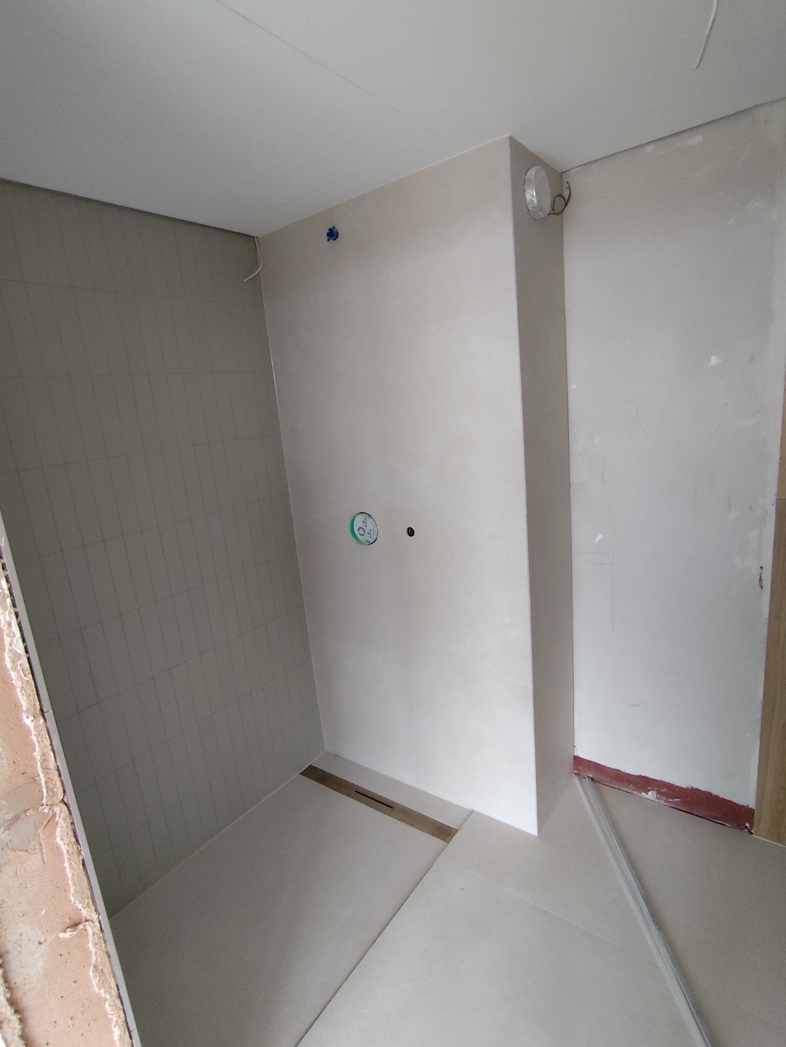 Wnętrze łazienki w trakcie wykańczania: ułożone płytki na podłodze i ścianach, widoczne instalacje. Minimalistyczny design, jasne kolory, surowy stan wykończenia.