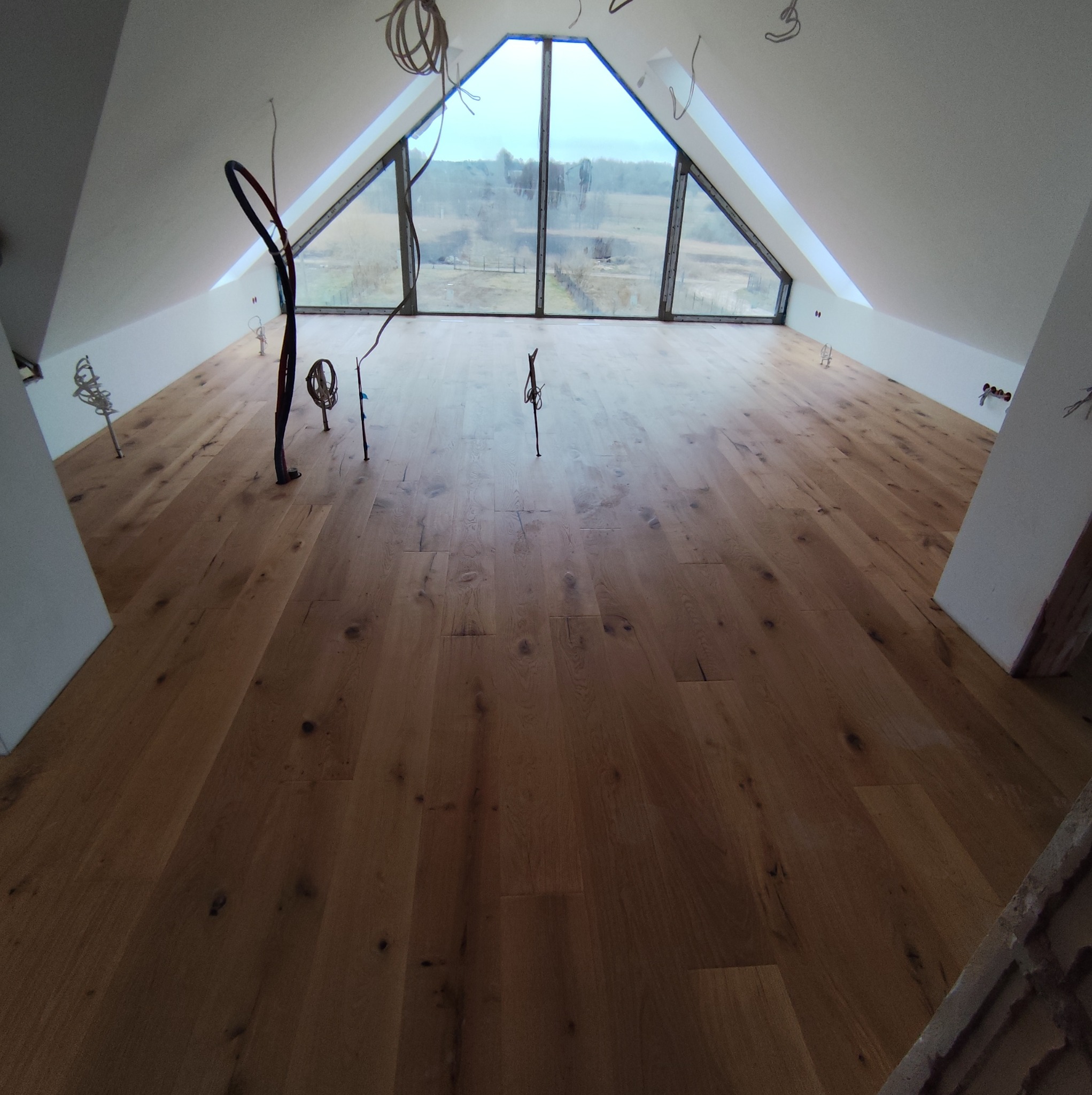 Jasna, drewniana podłoga w nowym poddaszu z oknem szczytowym. Widoczne kable elektryczne i fragmenty ścian w trakcie wykończenia. Naturalne światło dzienne.