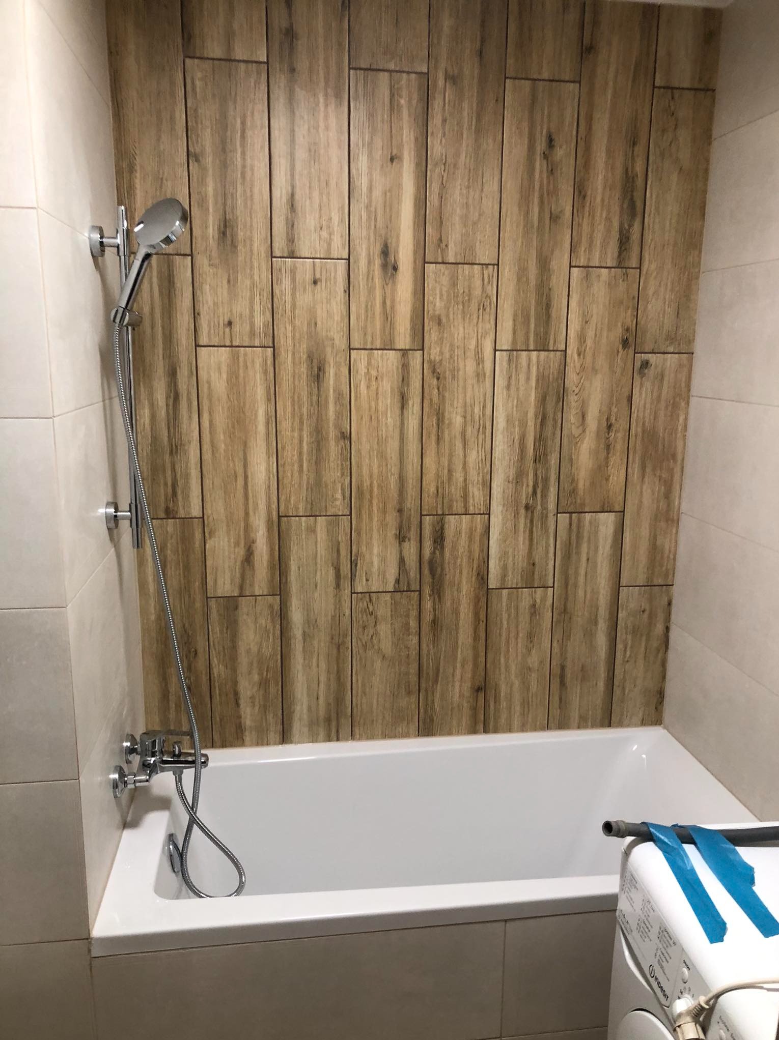 Wykończona łazienka z wanną i prysznicem, ściana wyłożona płytkami imitującymi drewno. Widoczna pralka z niebieską taśmą.