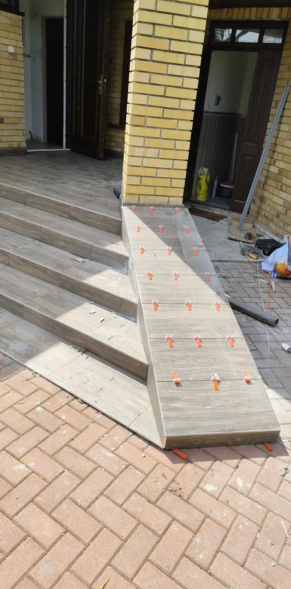 Kładzenie płytek na schodach wejściowych z użyciem klipsów poziomujących. Widoczne płytki imitujące drewno i ceglana elewacja budynku.