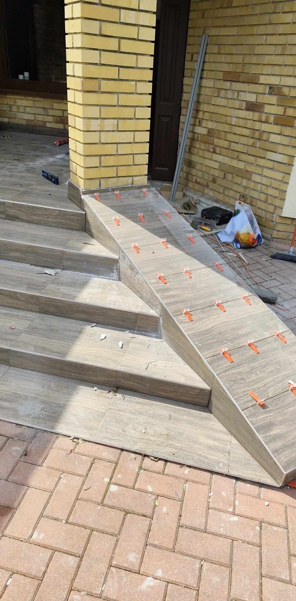 Układanie płytek na schodach wejściowych z rampą, z użyciem klipsów poziomujących, Białystok. Widoczny fragment ceglanej ściany i narzędzia pracy.