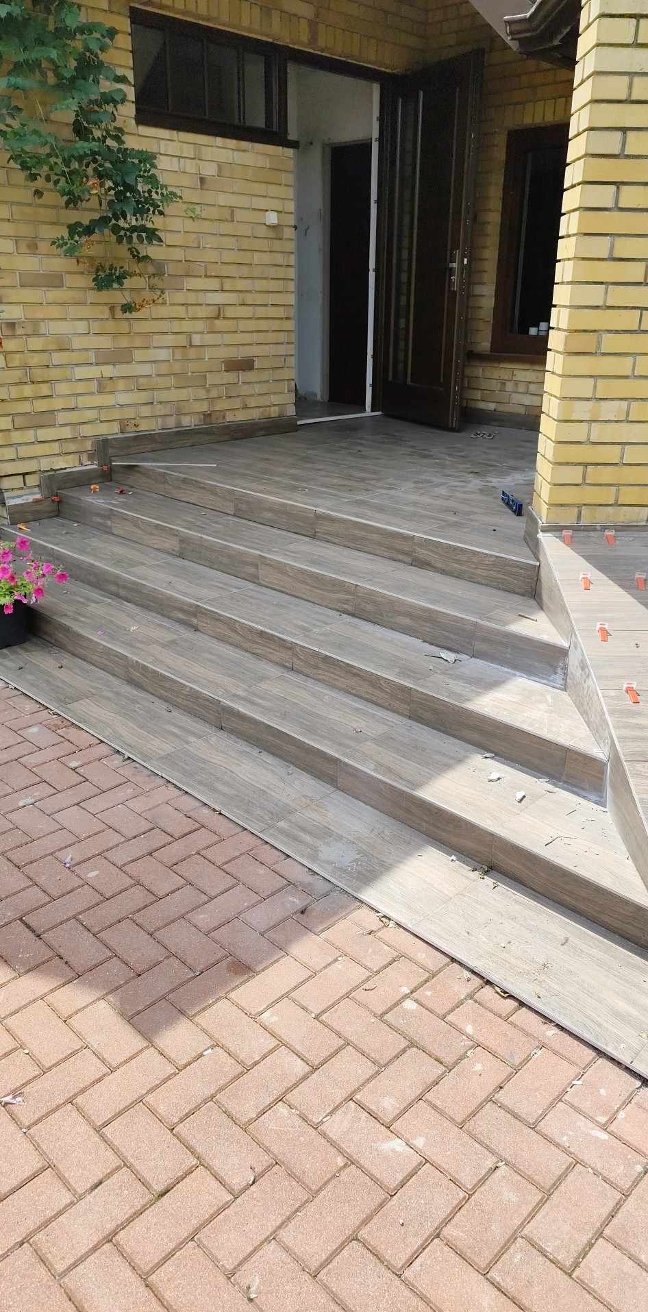 Wykończone schody wejściowe do domu, wyłożone płytkami imitującymi drewno, z widocznymi elementami mocującymi i fragmentem kostki brukowej. Otwarta, ciemna stolarka drzwiowa.