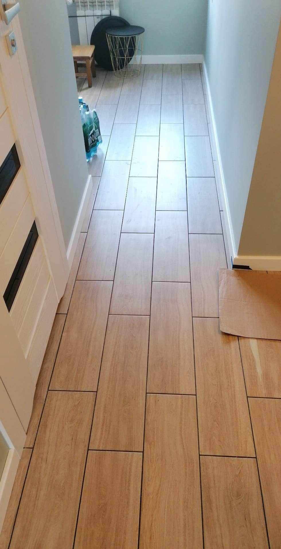 Ułożona podłoga z płytek imitujących drewno w korytarzu, widok z góry, fuga w kolorze czarnym, przy ścianie karton zabezpieczający podłogę.