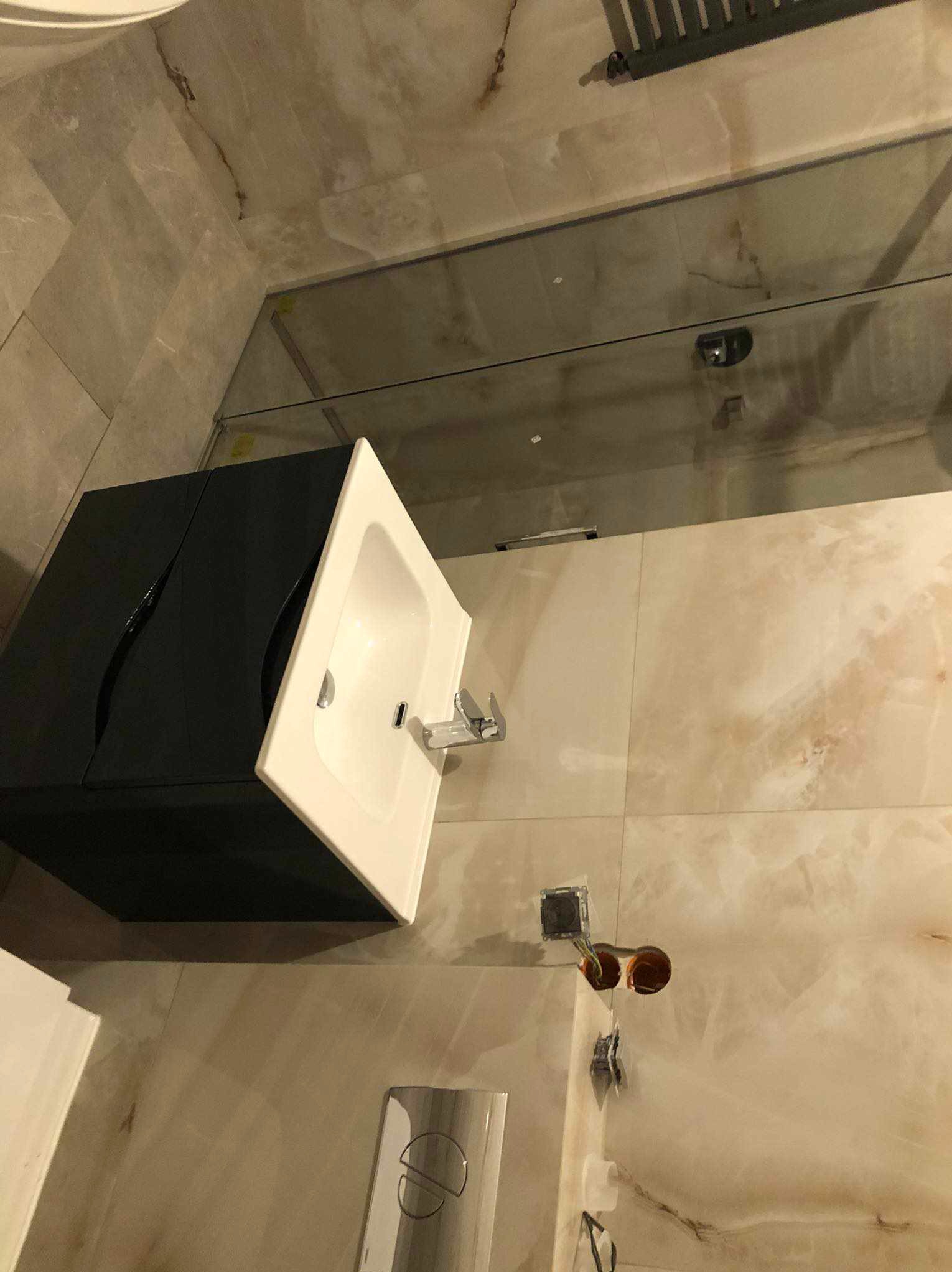 Nowoczesna łazienka po remoncie: czarna szafka z białą umywalką, beżowe płytki imitujące marmur, szklana kabina prysznicowa. Widoczne gniazdka elektryczne.