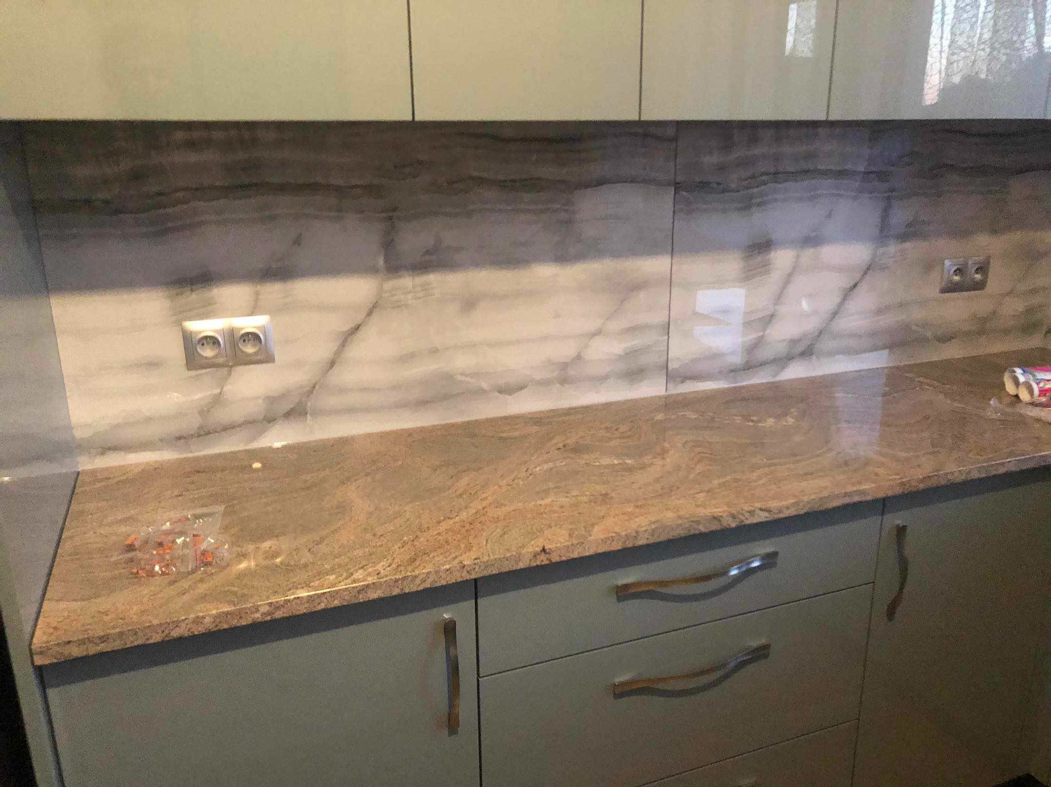 Wykończona kuchnia z marmurowym blatem i płytkami imitującymi marmur na ścianie. Widoczne gniazdka elektryczne i akcesoria montażowe na blacie.