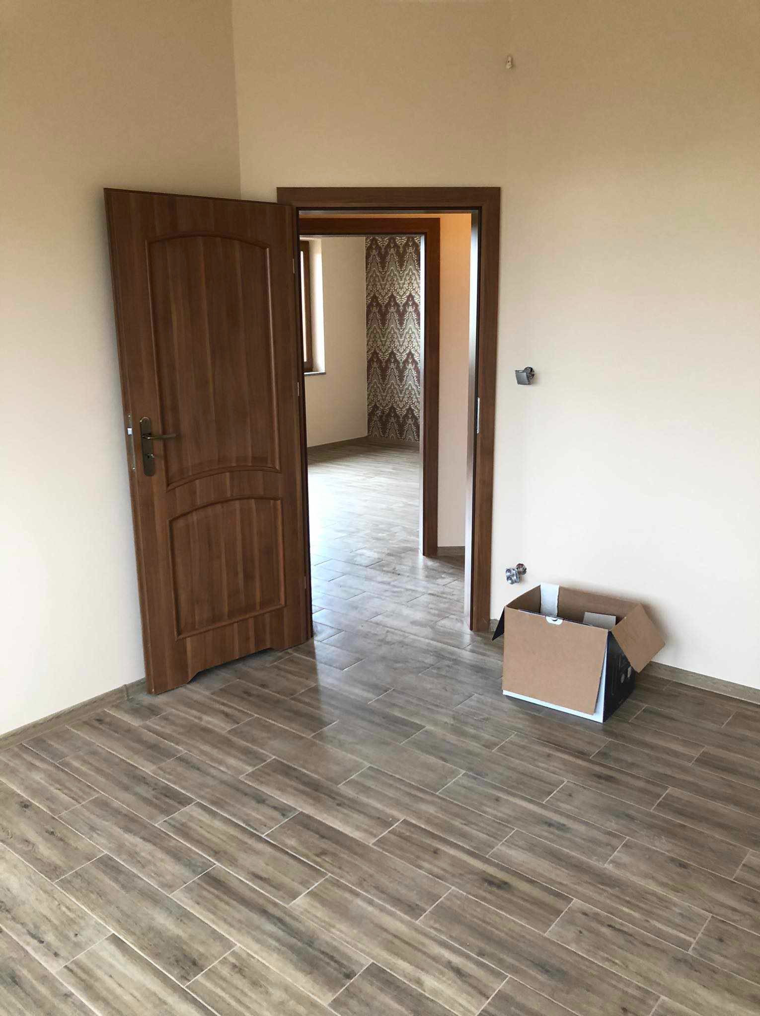 Wnętrze po remoncie: drewnopodobna podłoga z płytek, otwarte drewniane drzwi, karton na podłodze, widok na pokój z wzorzystą tapetą. Jasne ściany.