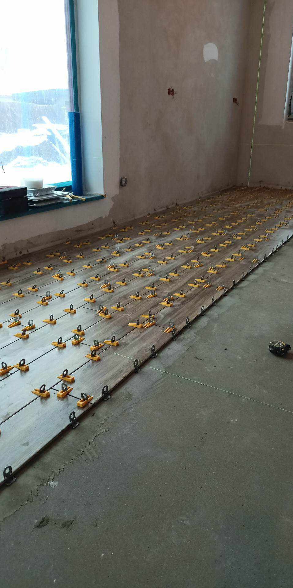 Układanie płytek podłogowych z użyciem systemu poziomowania, widoczny laser krzyżowy. Surowe ściany w tle, okno z niebieską folią zabezpieczającą.