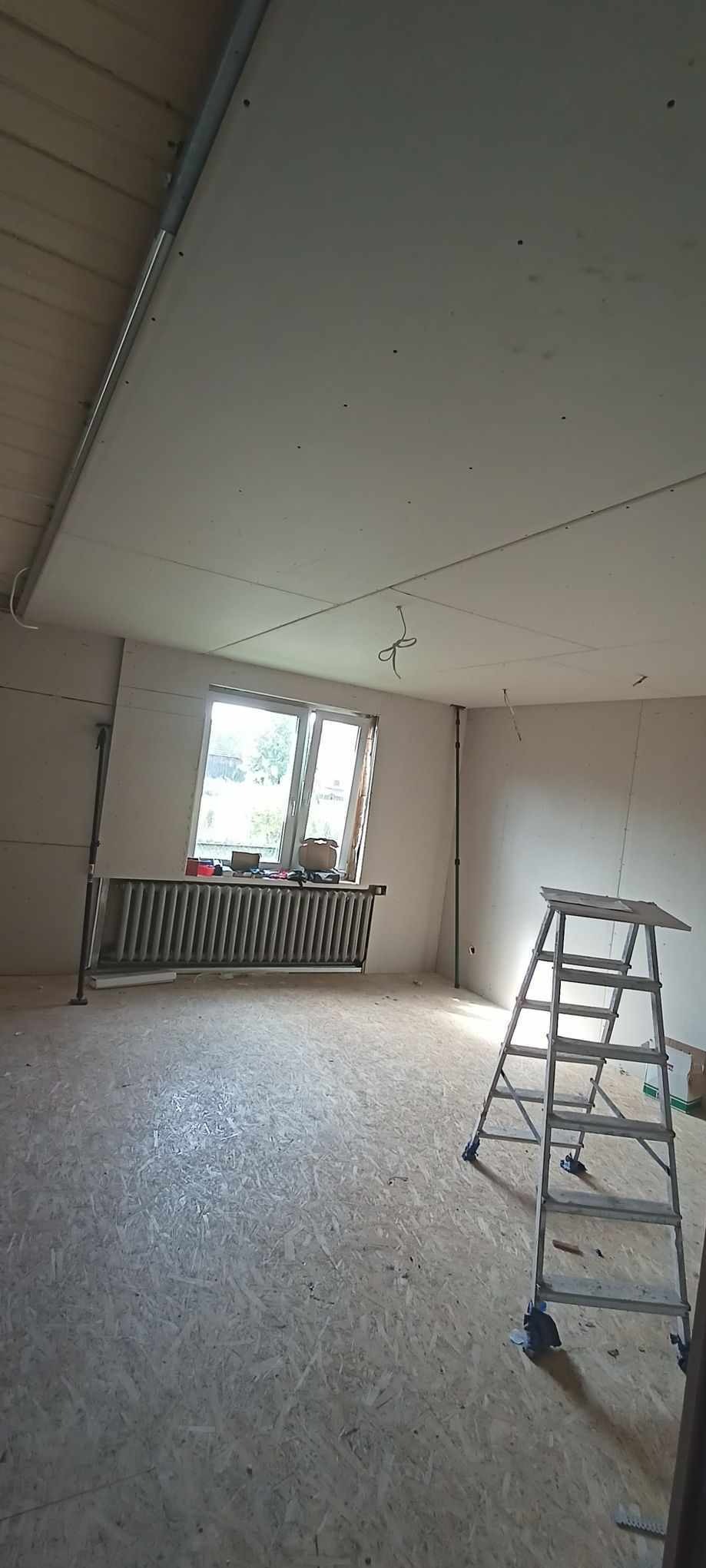 Pokój w trakcie remontu: zamontowana płyta karton-gips na suficie, widoczna drabina i grzejnik pod oknem. Podłoga wyłożona płytami OSB. Prace wykończeniowe w toku.