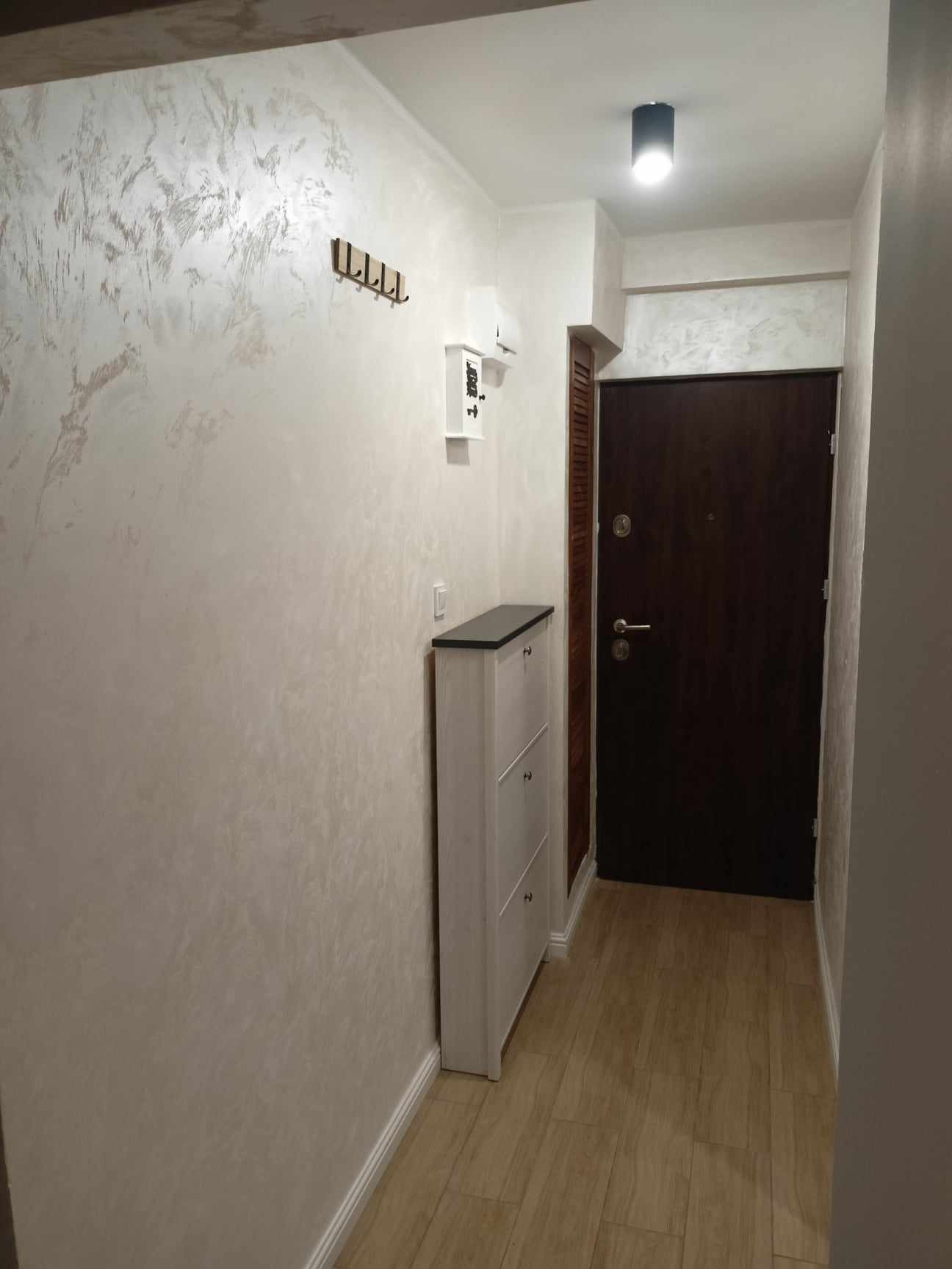 Wykończony korytarz z dekoracyjną ścianą w odcieniach beżu, drewnianą podłogą, szafką na buty i ciemnymi drzwiami wejściowymi. Widoczne wieszaki i oświetlenie sufitowe.