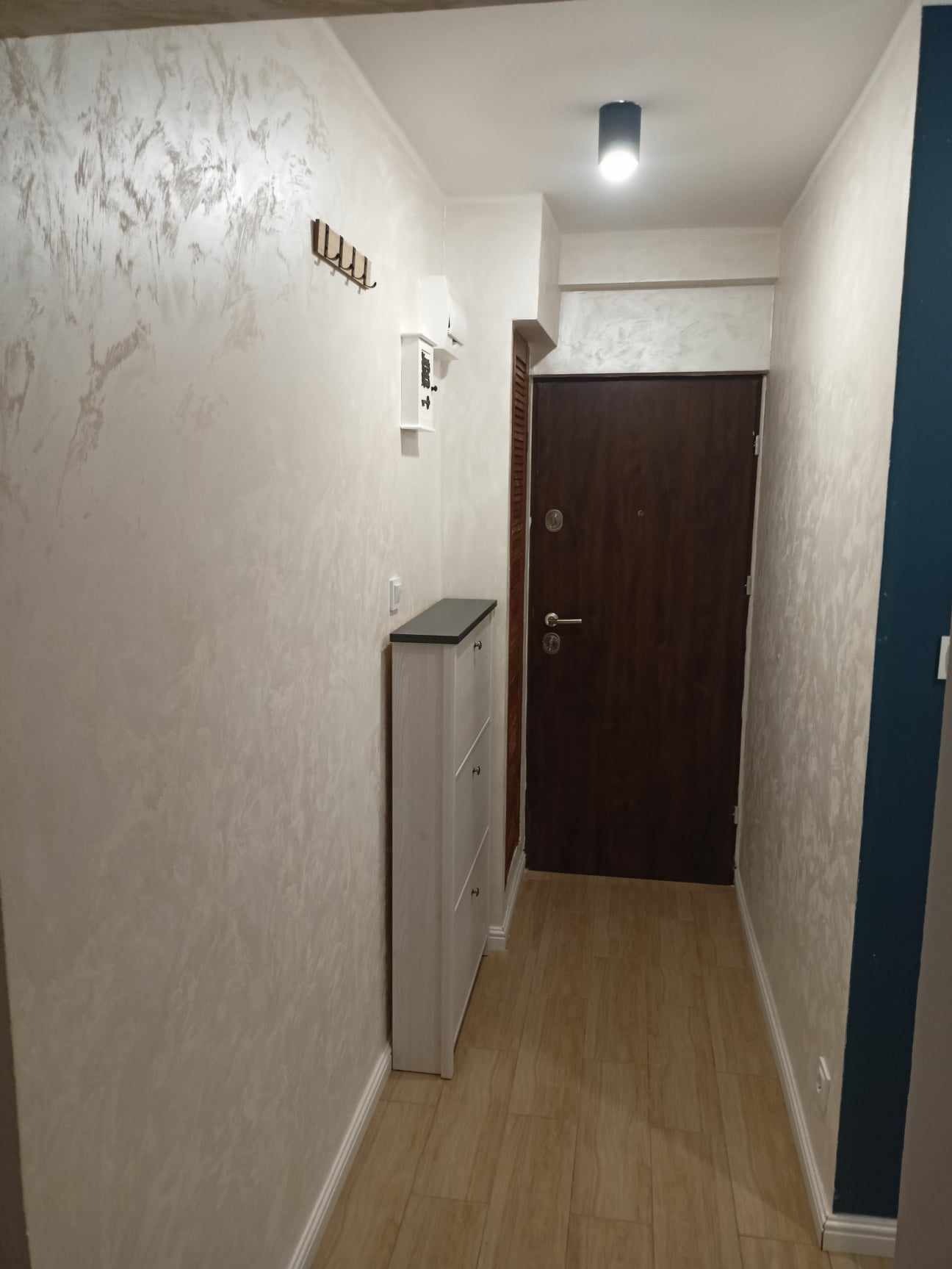 Wykończony korytarz z drewnianą podłogą, szafką na buty i wieszakami na ścianie. Ściany pokryte dekoracyjnym tynkiem w odcieniach bieli i szarości. Ciemne drzwi wejściowe.