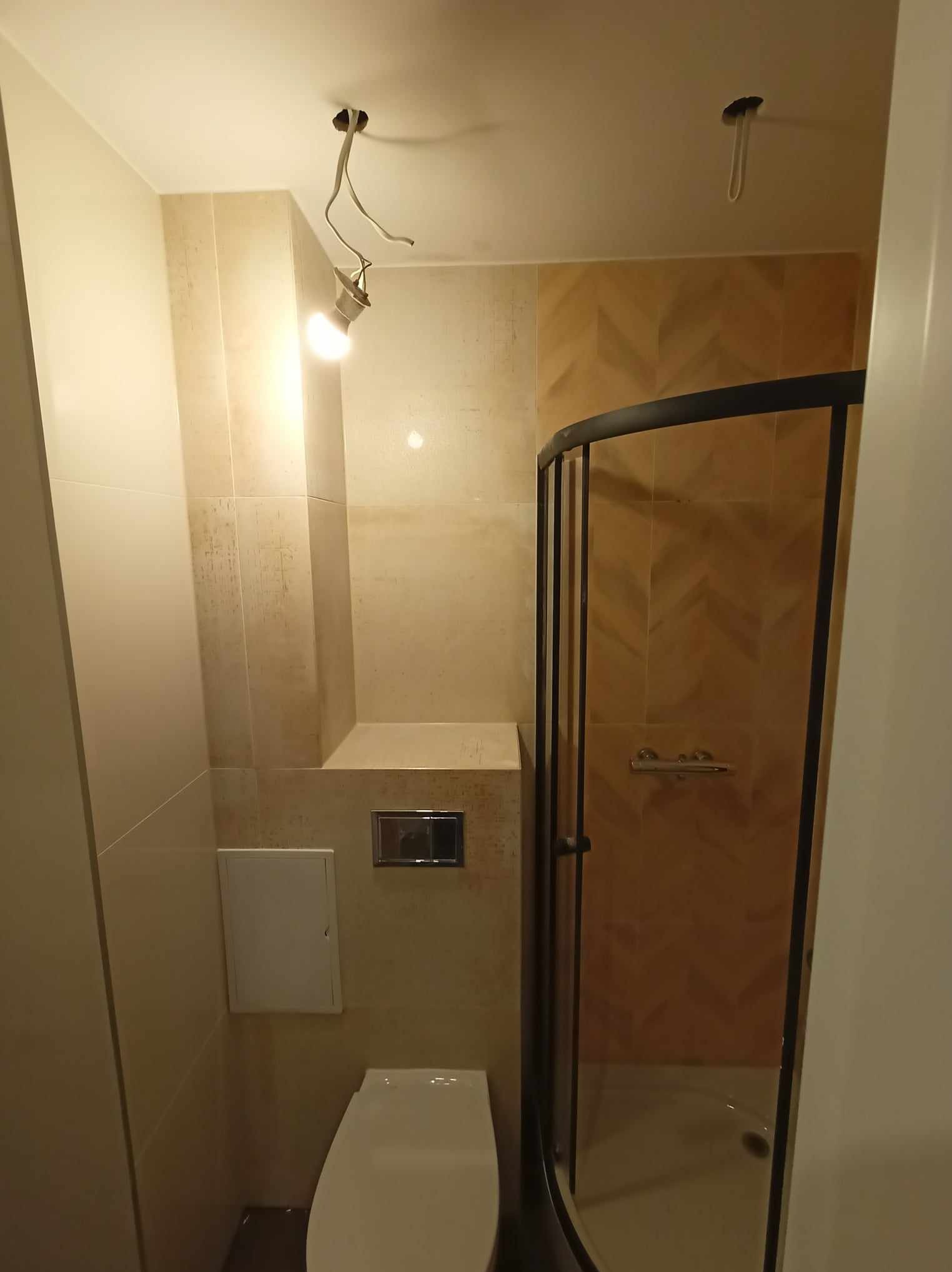 Beżowa łazienka po remoncie: kabina prysznicowa z czarną ramą, podwieszana toaleta, płytki imitujące drewno na ścianach, surowa żarówka. Minimalistyczny design.